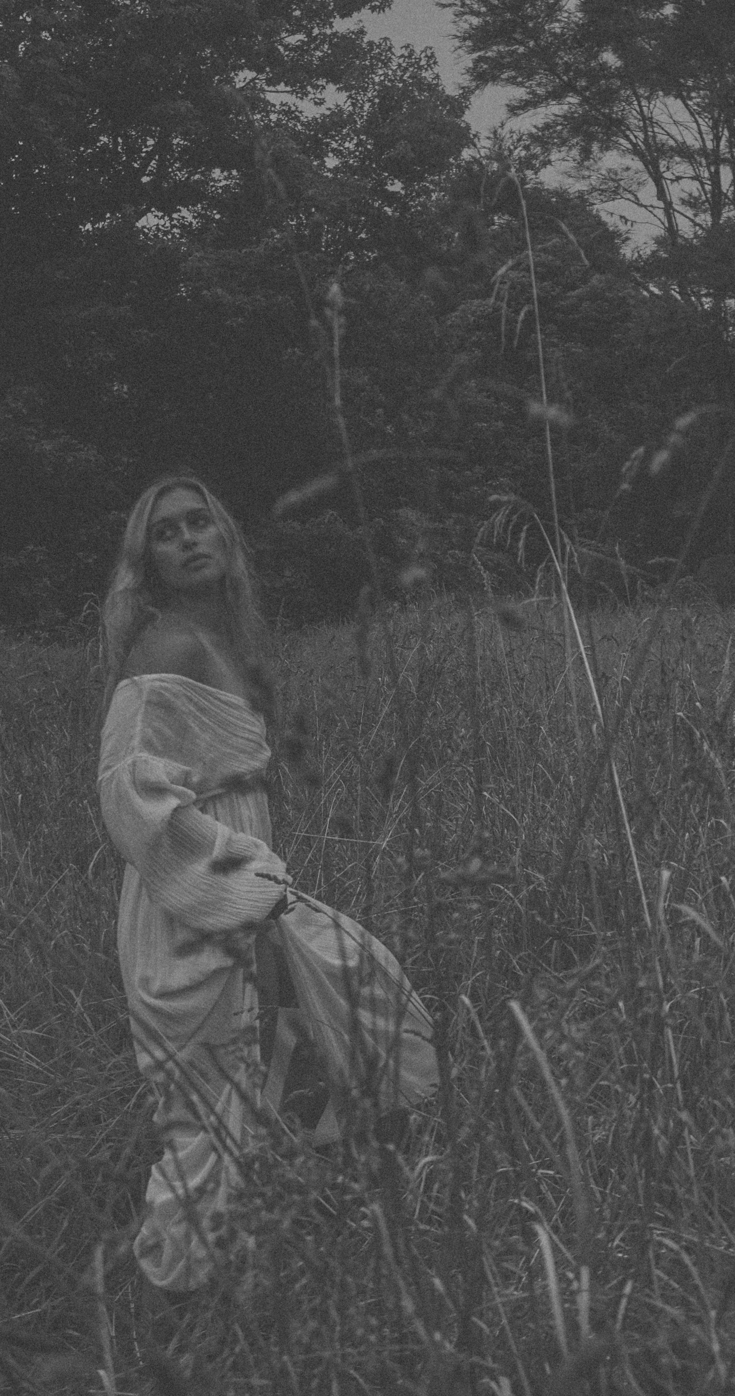 Black and white artistic portrait in tall grass with dramatic lighting and flowing fabric.