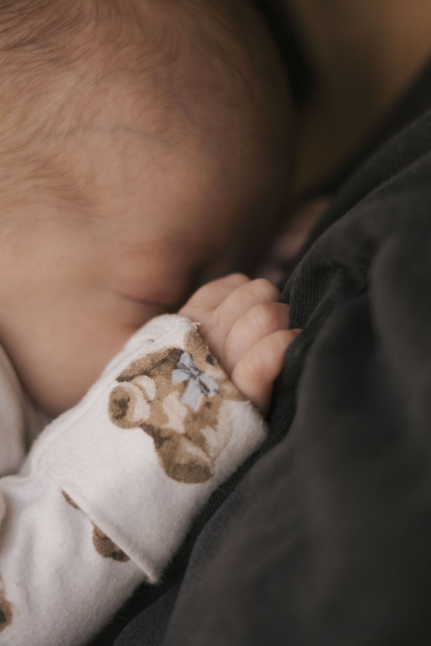 Close-up of sleeping baby wearing patterned white onesie against dark fabric.