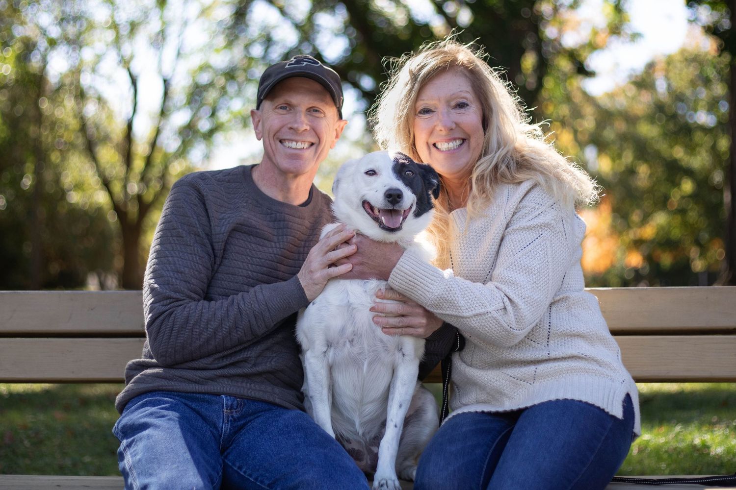 A couple sits on a bench outdoors with their happy white fluffy dog on a sunny day. Pet photography in Burlington, Iowa