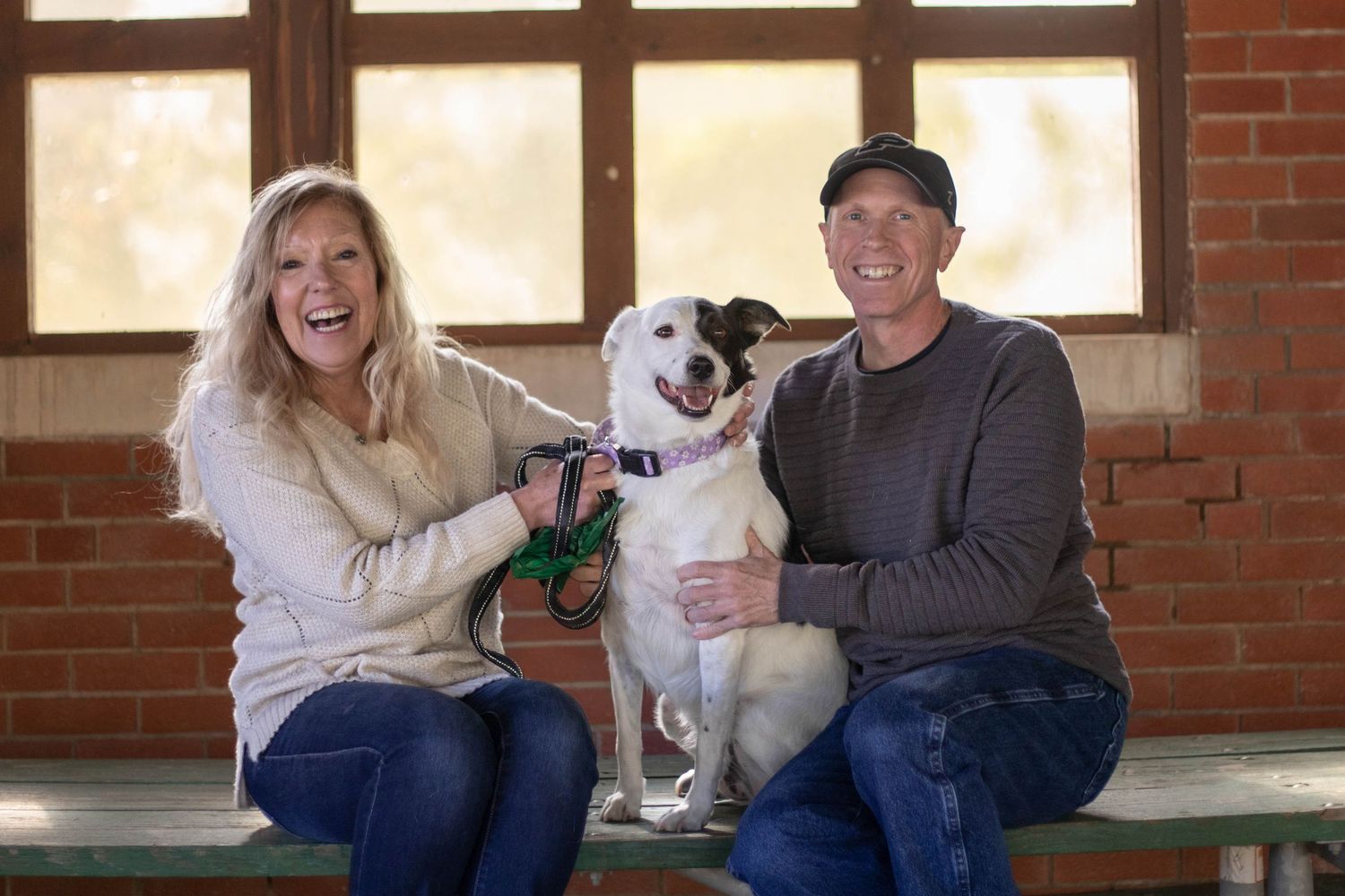 A group of people sit on a bench outdoors while laughing and playing with a white dog. Pet photography in Burlington, Iowa