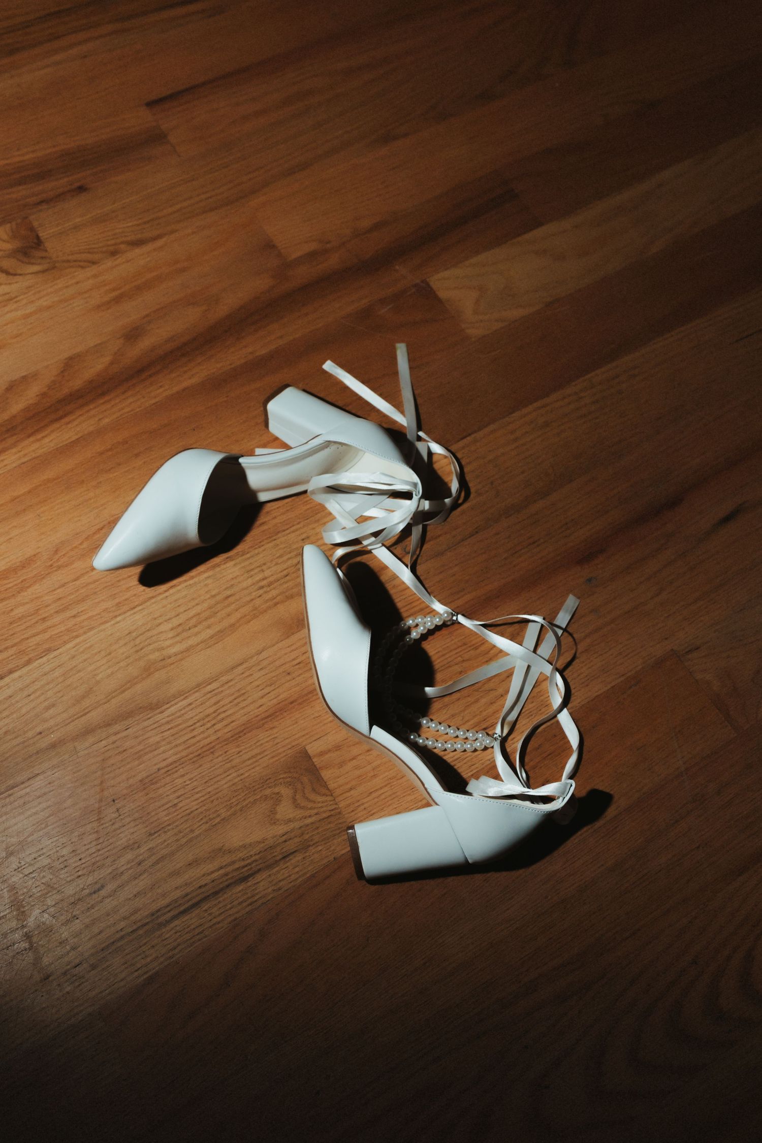 White strappy wedding heels arranged artistically on a wooden floor with dramatic lighting.
