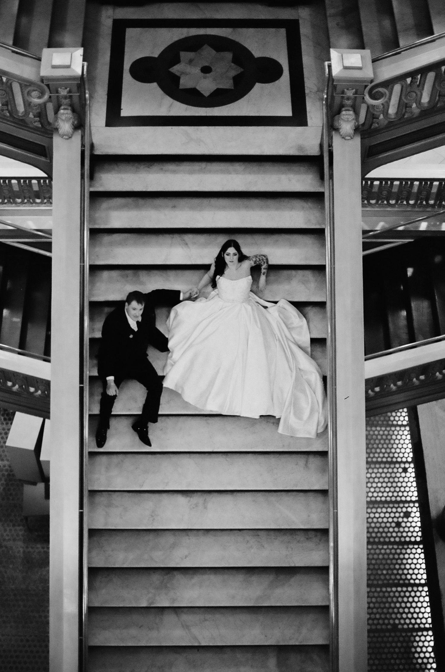 Black and white photo of bride and groom sitting on grand marble staircase, shot from above on wedding day.