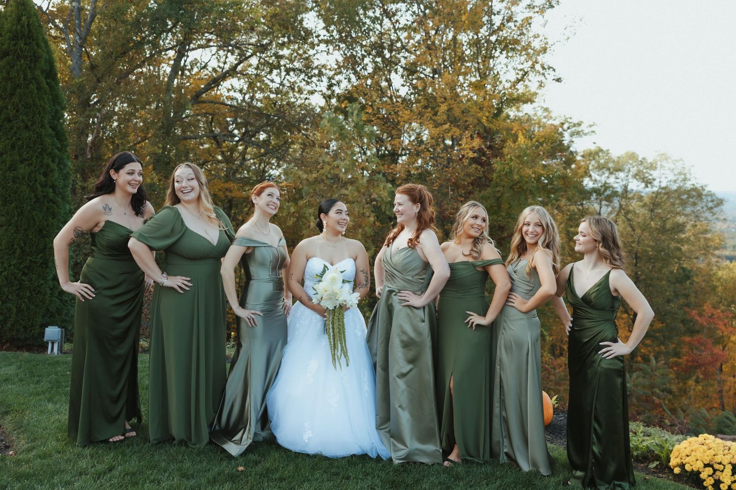 Wedding party in sage green dresses poses together outdoors during autumn with colorful fall foliage in background.