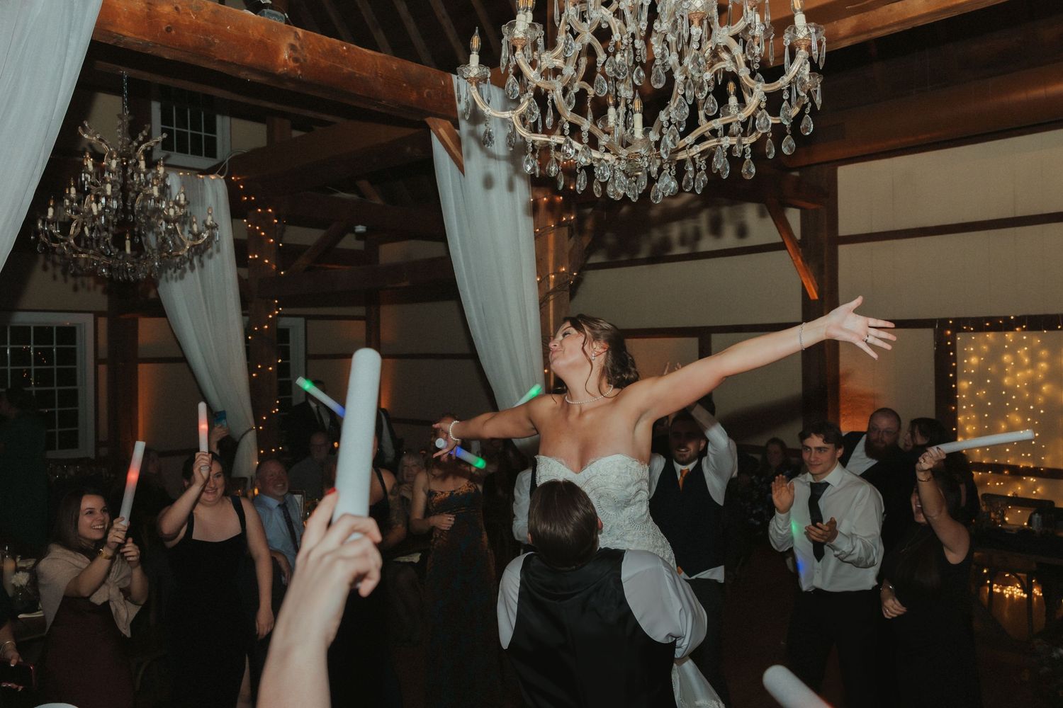 Wedding guests dance and celebrate with arms raised under a crystal chandelier at an indoor reception venue.