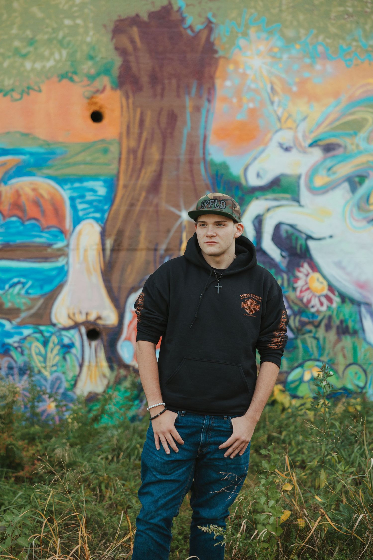 Person in black hoodie and baseball cap poses in front of colorful graffiti mural featuring trees and fantasy elements.