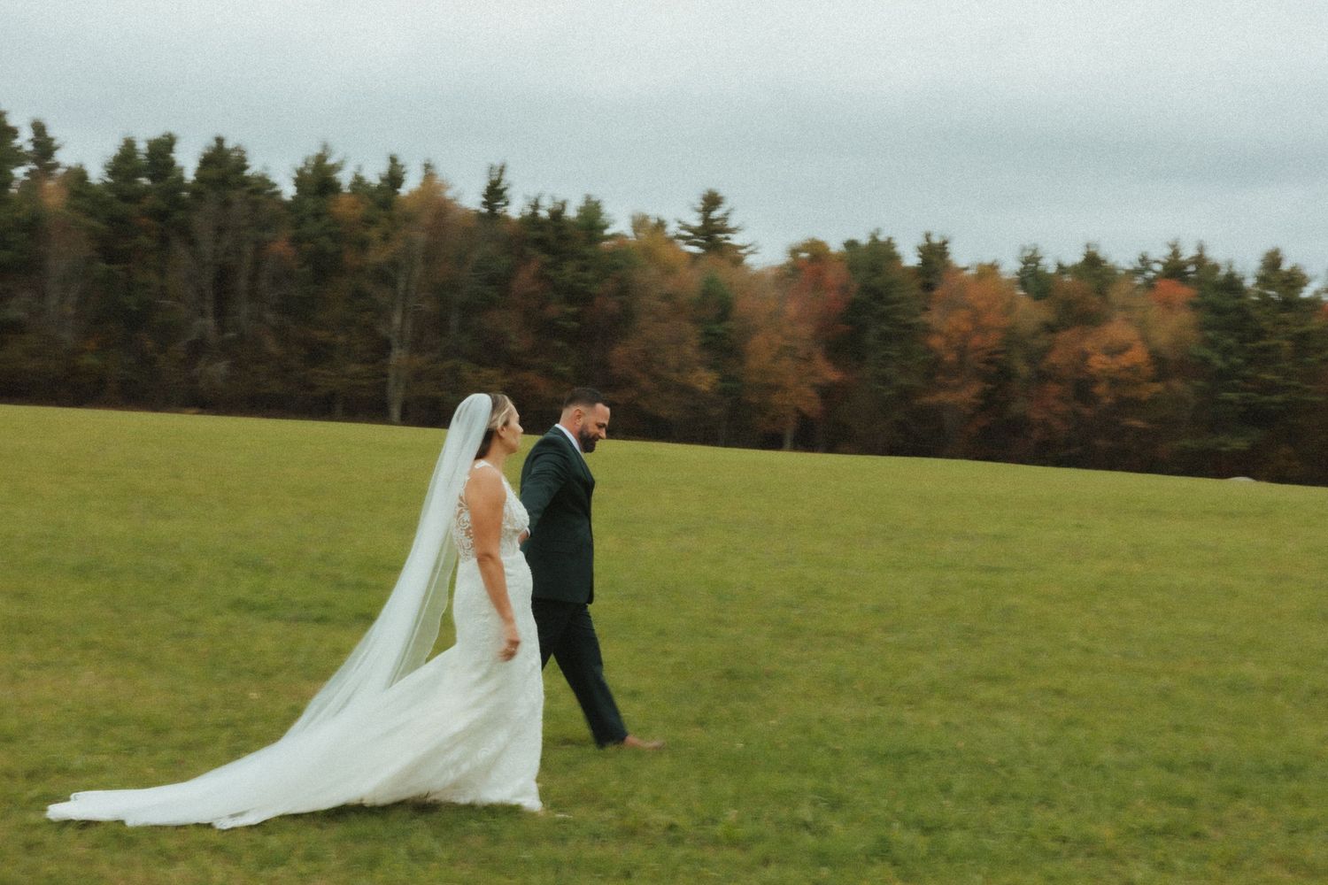 A couple in wedding attire walks across a green field with autumn trees in the background during their wedding photoshoot.
