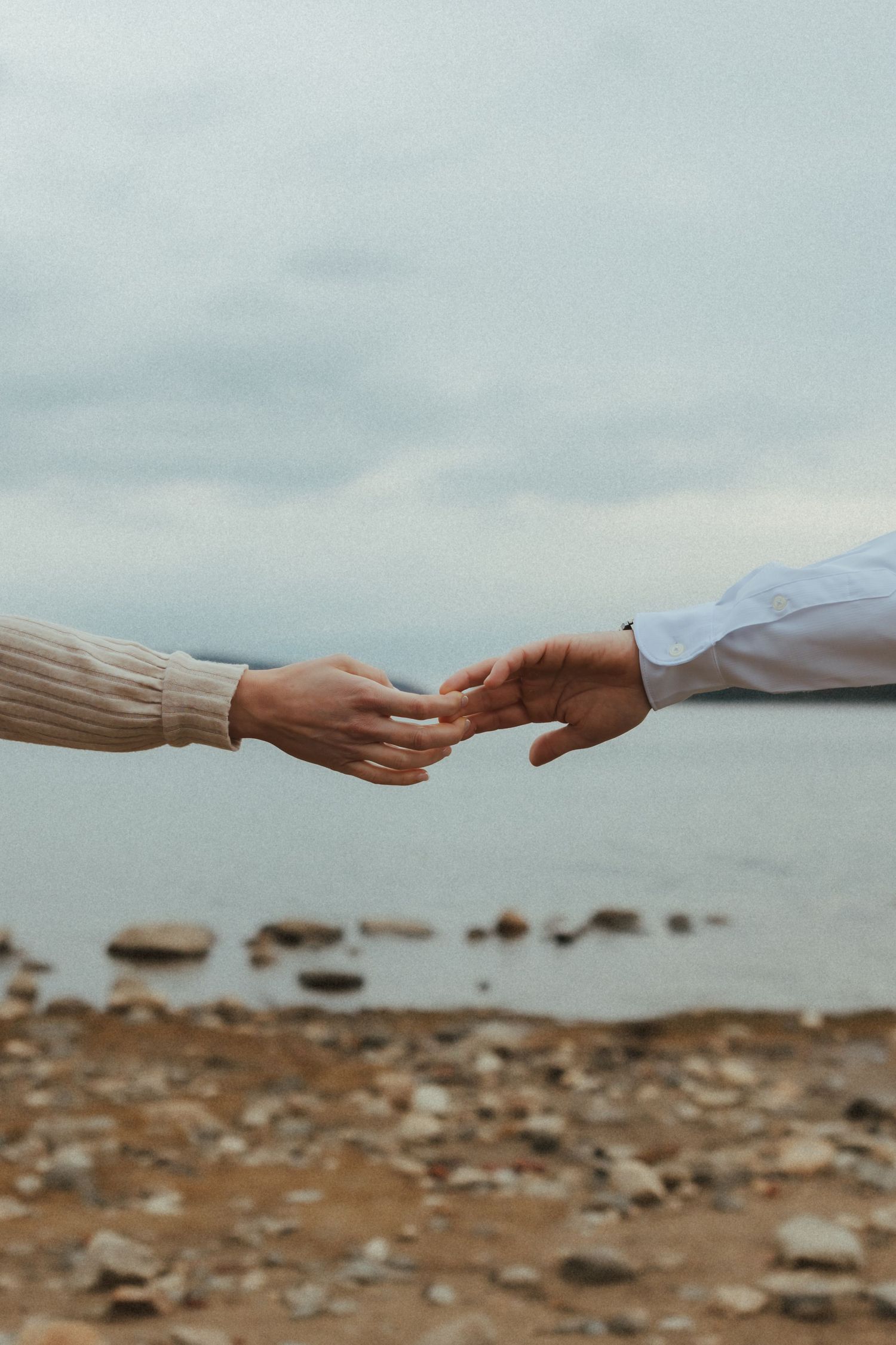 Two hands reaching towards each other against a rocky beach landscape with cloudy skies in the background.