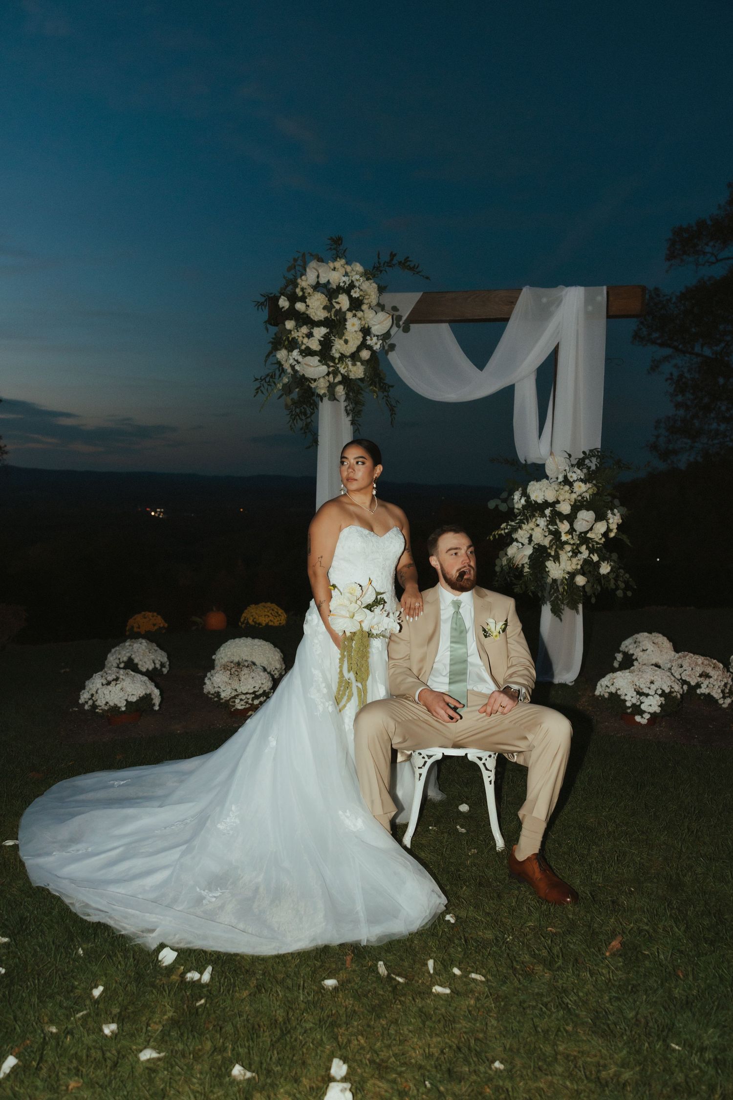 A couple in wedding attire sits together on grass at dusk with white floral decorations and flowing fabric nearby.