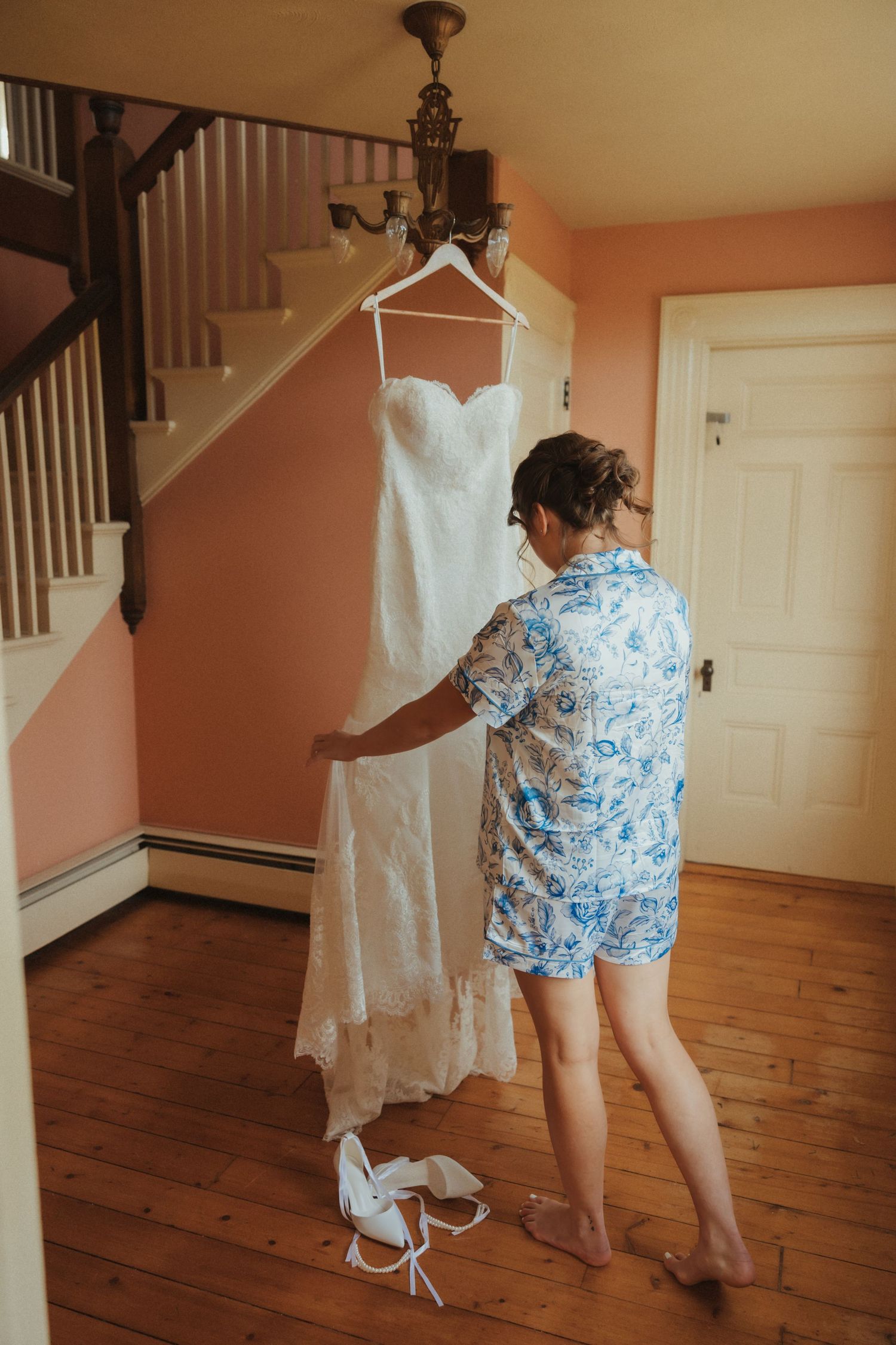 A wedding dress hangs in a warm-lit room while someone in blue floral shorts examines the details.