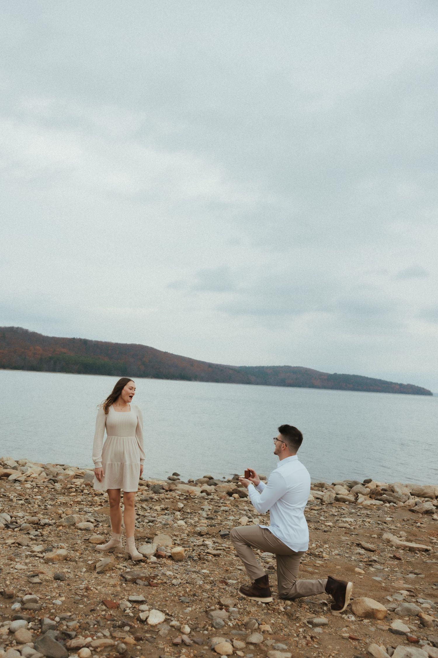 A couple shares a romantic moment on a rocky shoreline overlooking a misty lake and mountains.