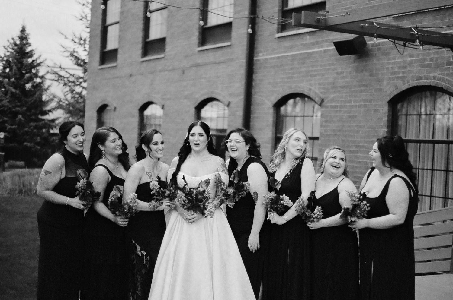 Black and white photo of a bride in a white gown surrounded by bridesmaids in black dresses holding bouquets.