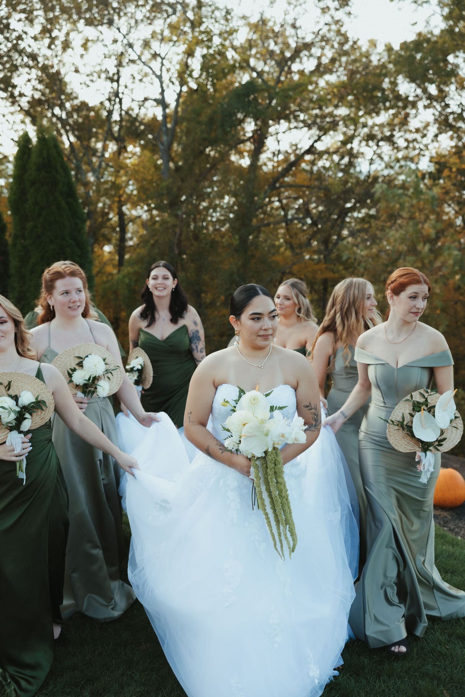 Wedding party in sage green bridesmaid dresses poses outdoors with white bouquets during autumn sunset.