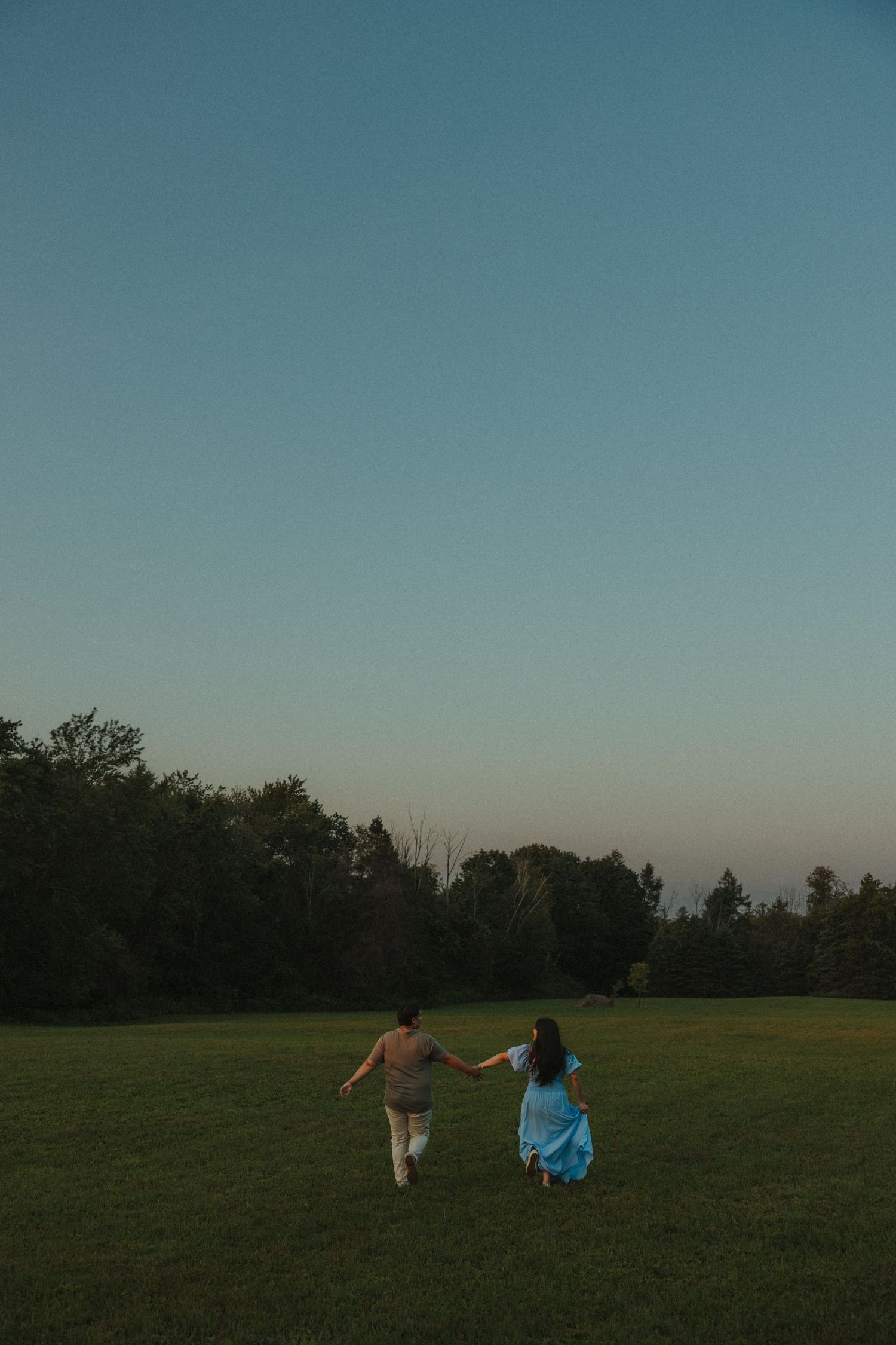 Two people sit together on a grassy field at dusk with trees visible in the background silhouette.
