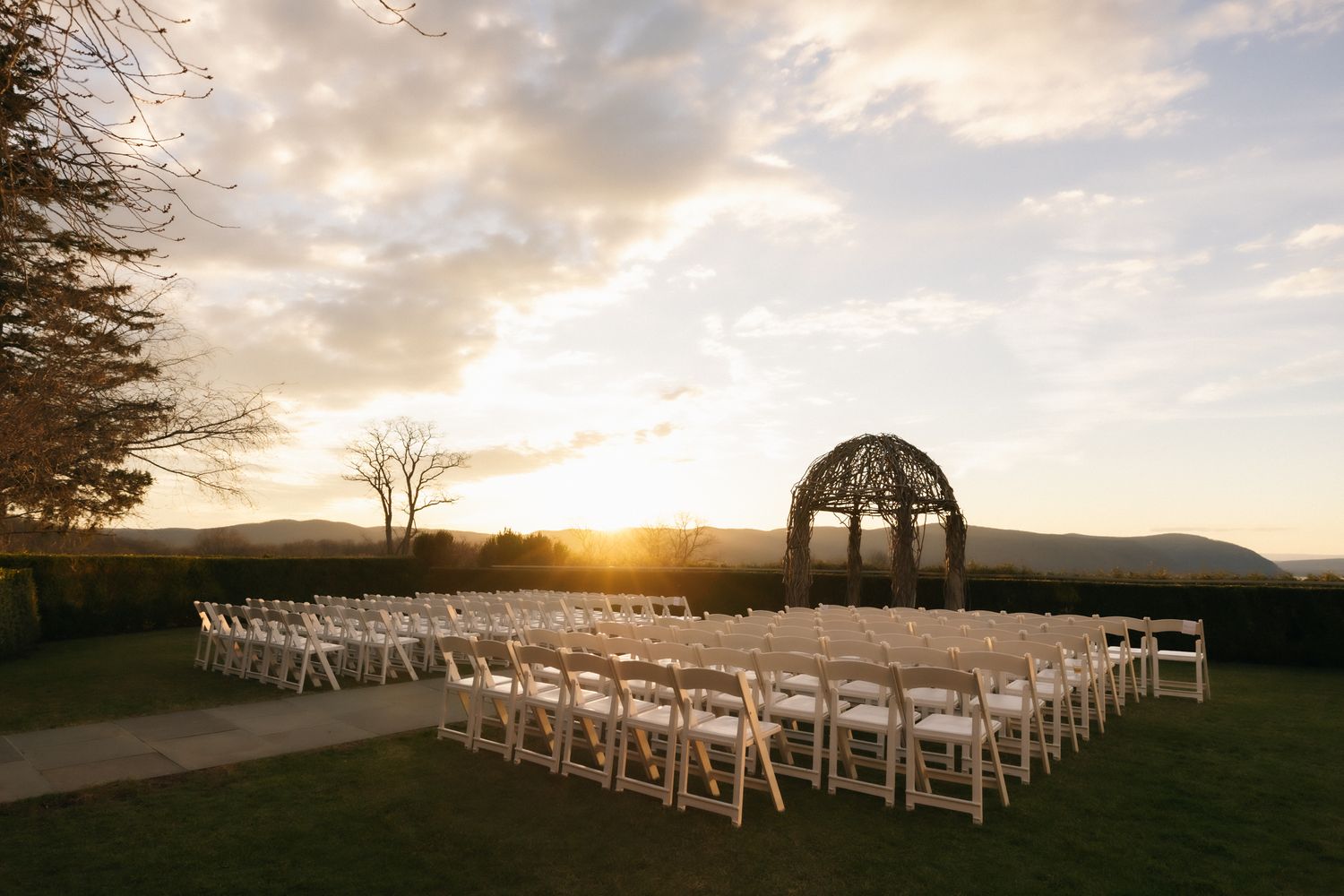 Outdoor wedding ceremony setup with white chairs and rustic arch at golden sunset with mountain views.