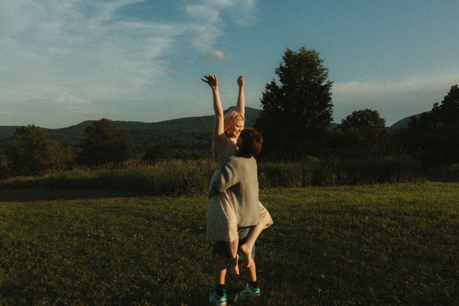 A couple embraces in a sunlit meadow during golden hour with mountains and trees in the background.