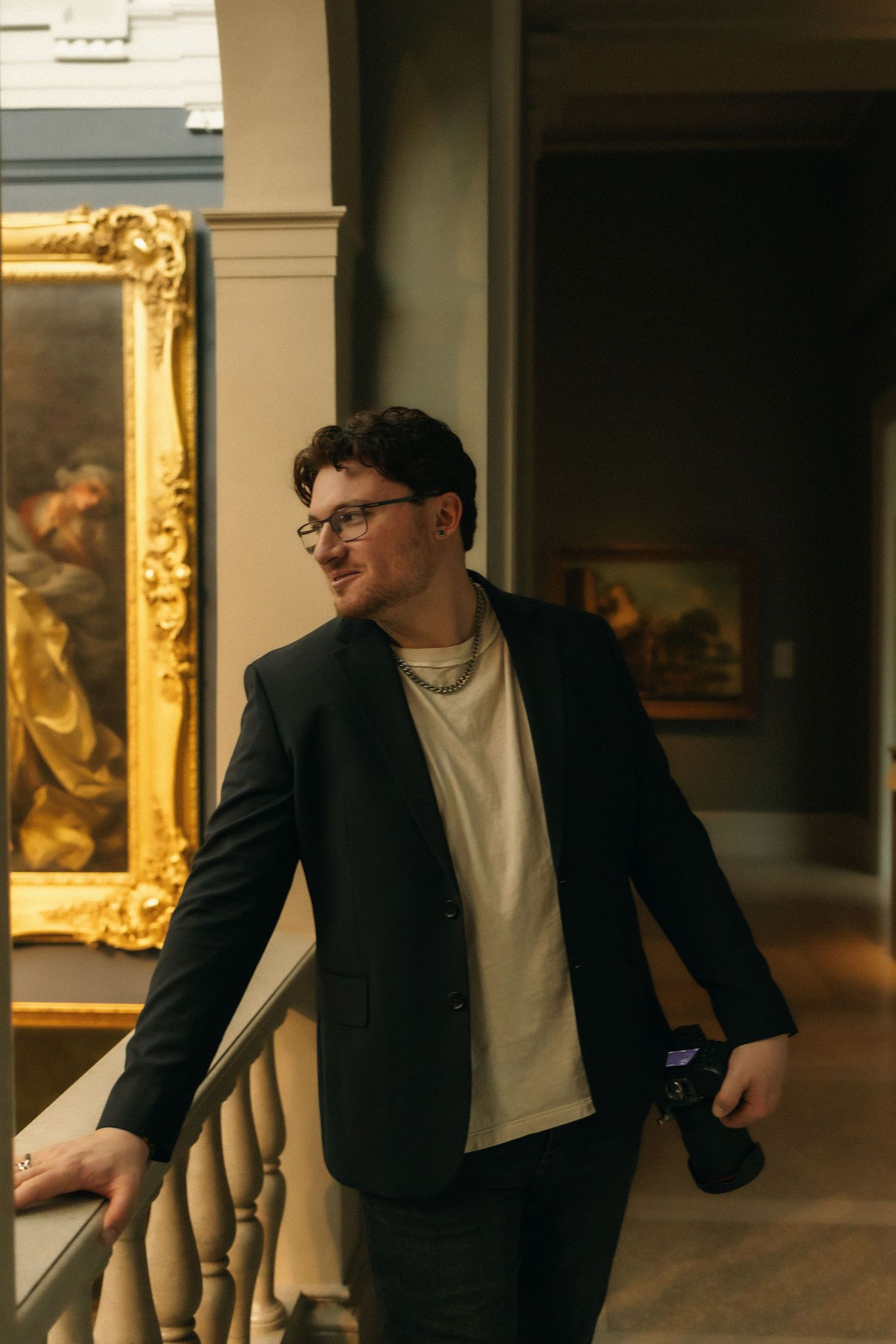 Man in black blazer holding camera smiles in art museum gallery with gold-framed paintings.