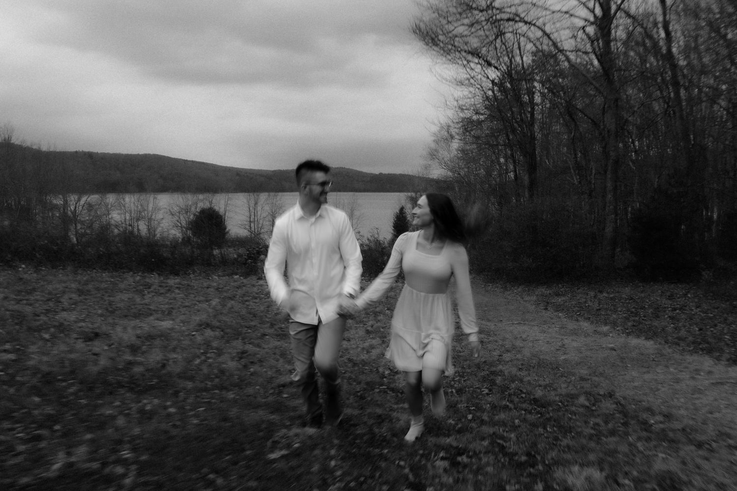 A couple in white outfits runs playfully through a field on an overcast day.