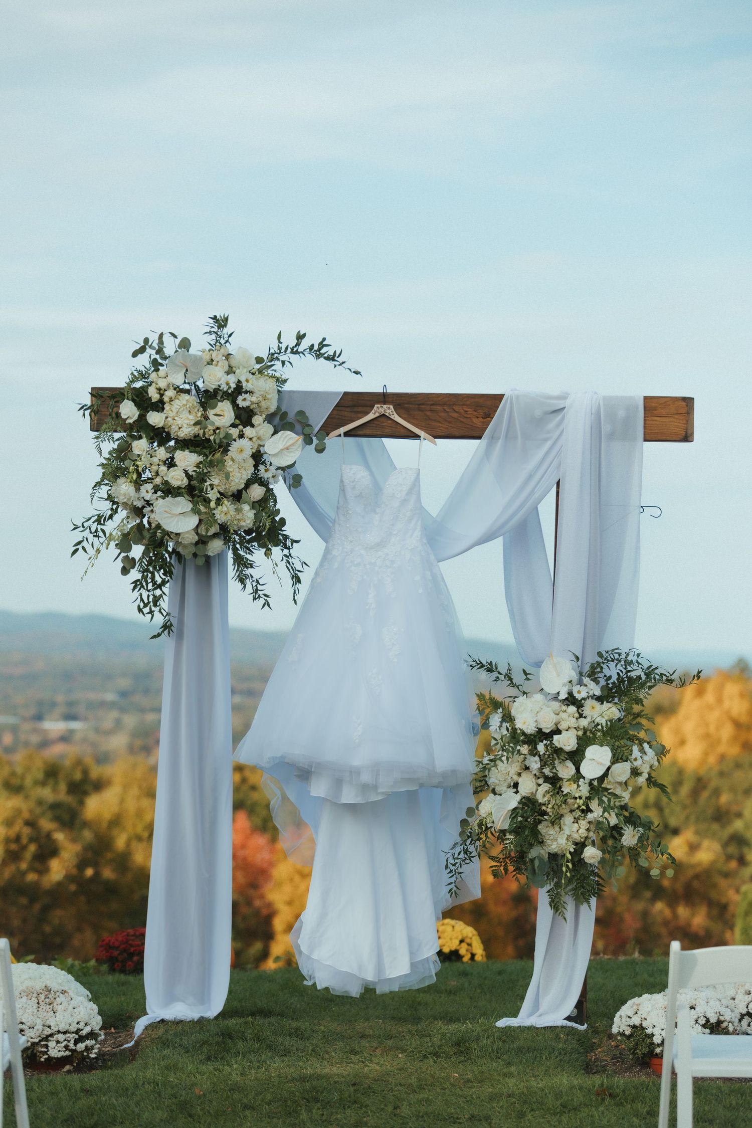 Elegant white wedding arch decorated with white roses and flowing fabric overlooks a scenic autumn landscape.