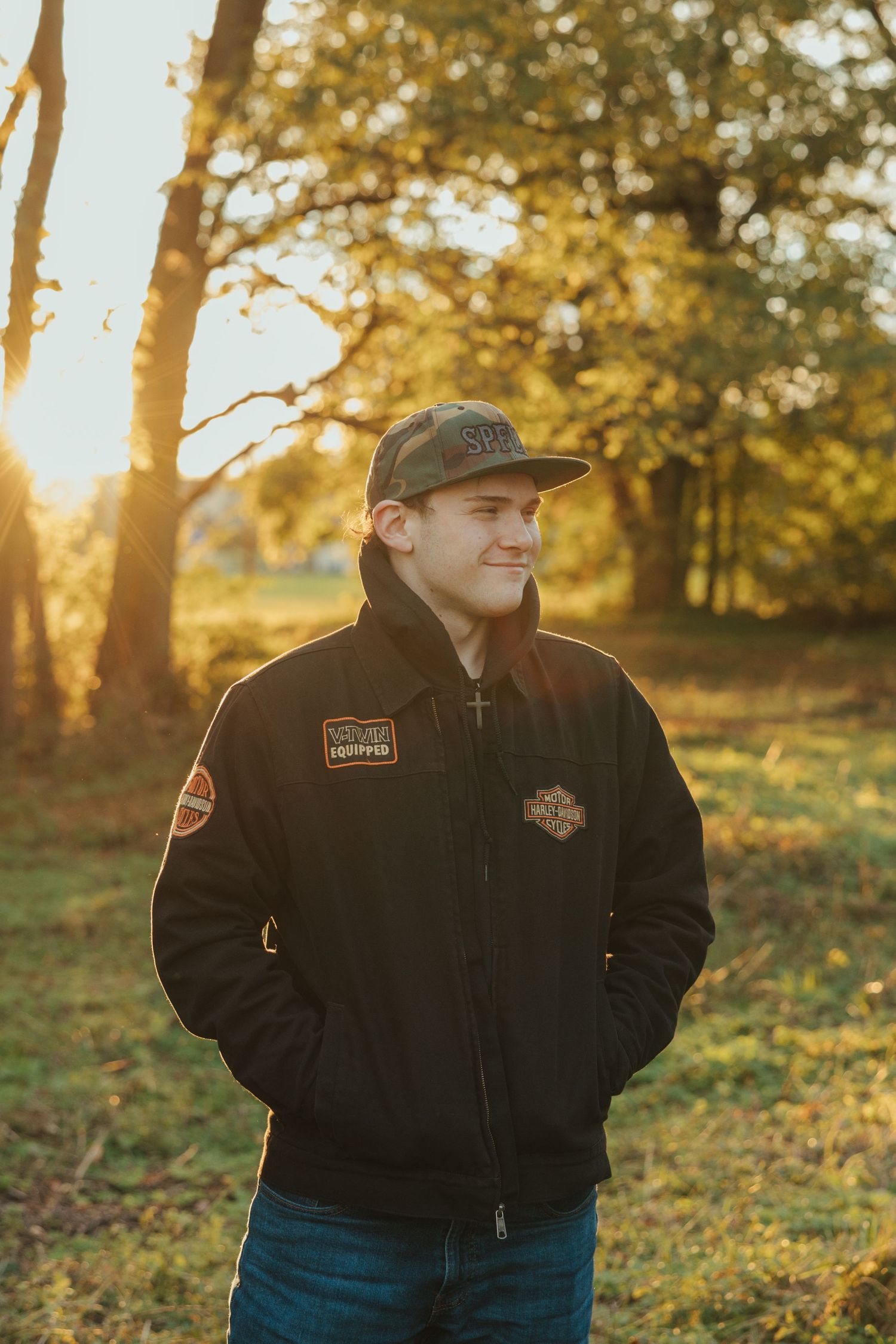 Person in black jacket and baseball cap stands outdoors during golden hour sunset with autumn trees in background.