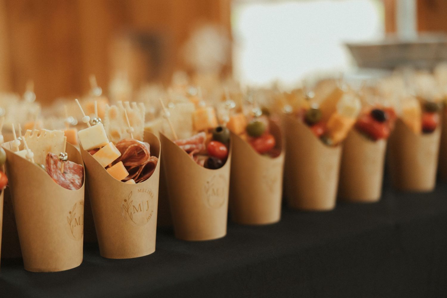 Brown paper cones filled with food are arranged in a line on a dark surface at a catering event.