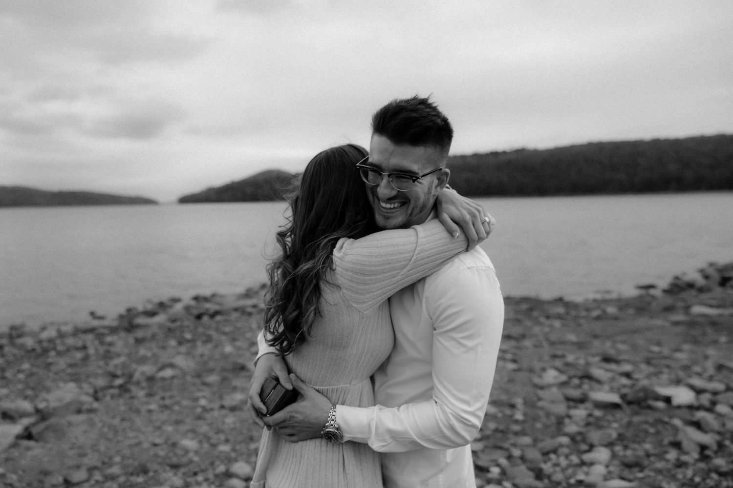 A romantic couple embraces on a rocky shoreline with mountains and water in the background in black and white.
