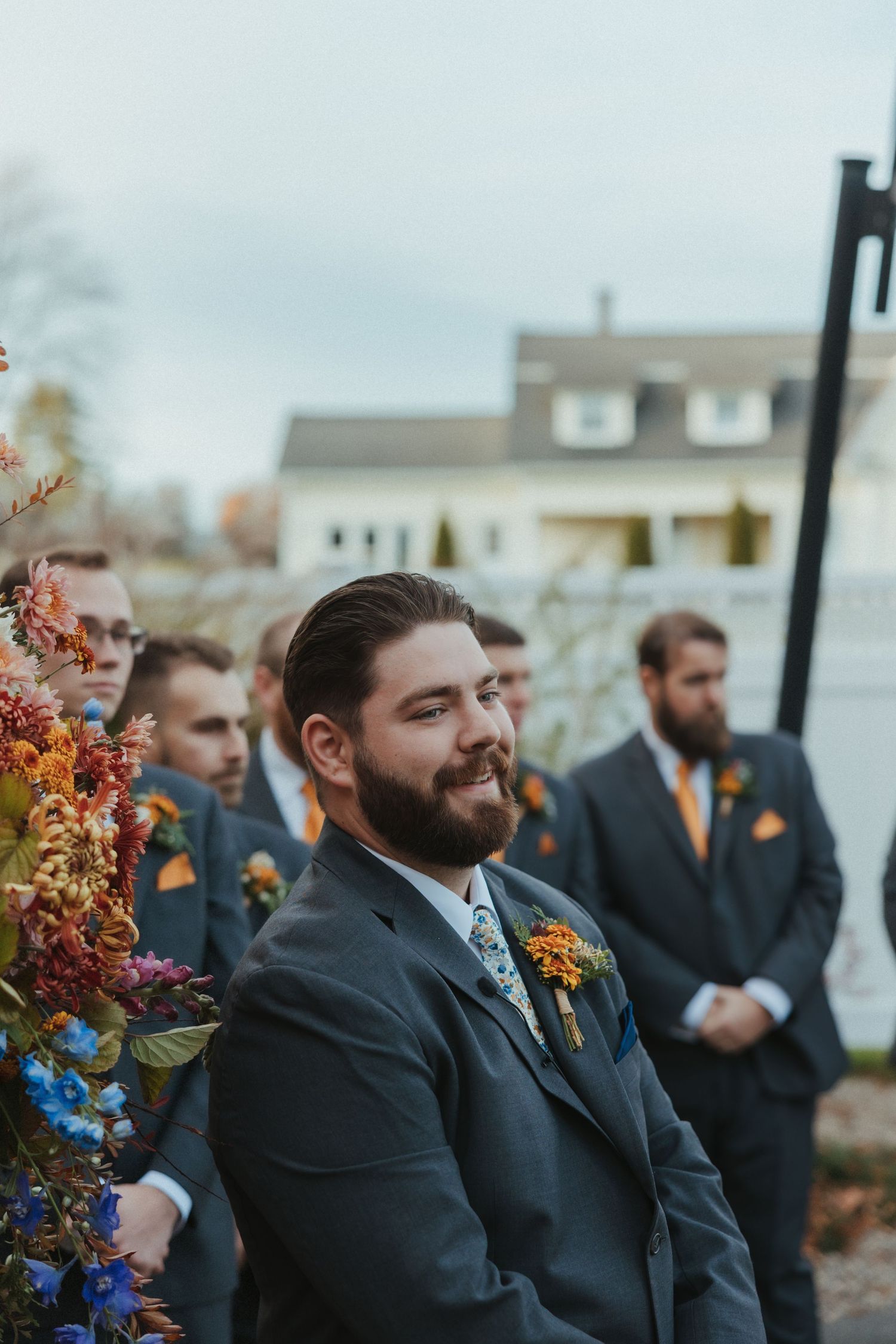 Groomsmen in dark suits with orange boutonnieres smile during an outdoor autumn wedding ceremony.