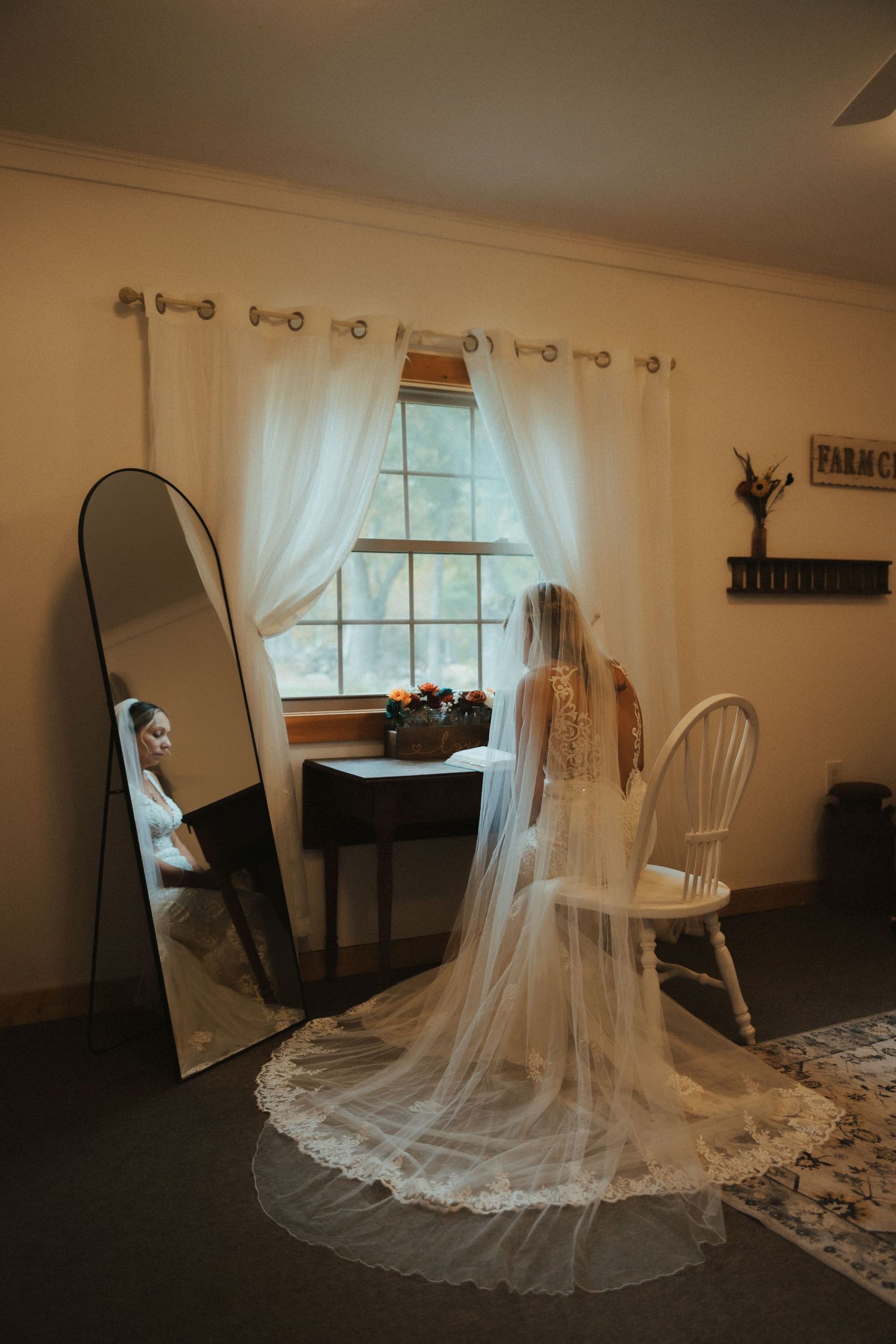 Wedding dress with long lace veil displayed in a room with a full-length mirror and white curtained window.