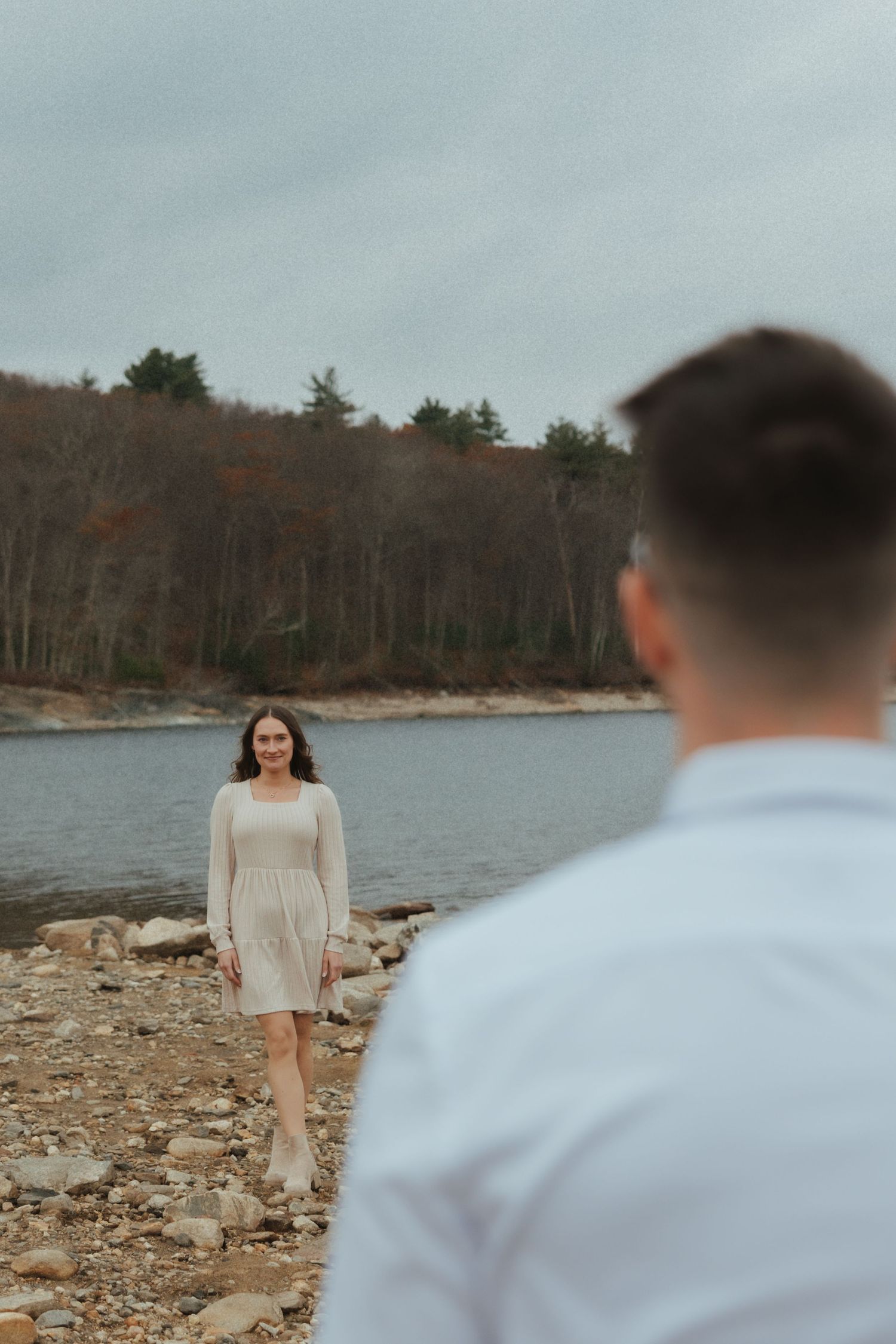 A couple takes romantic engagement photos on a rocky beach shoreline during an overcast autumn day.