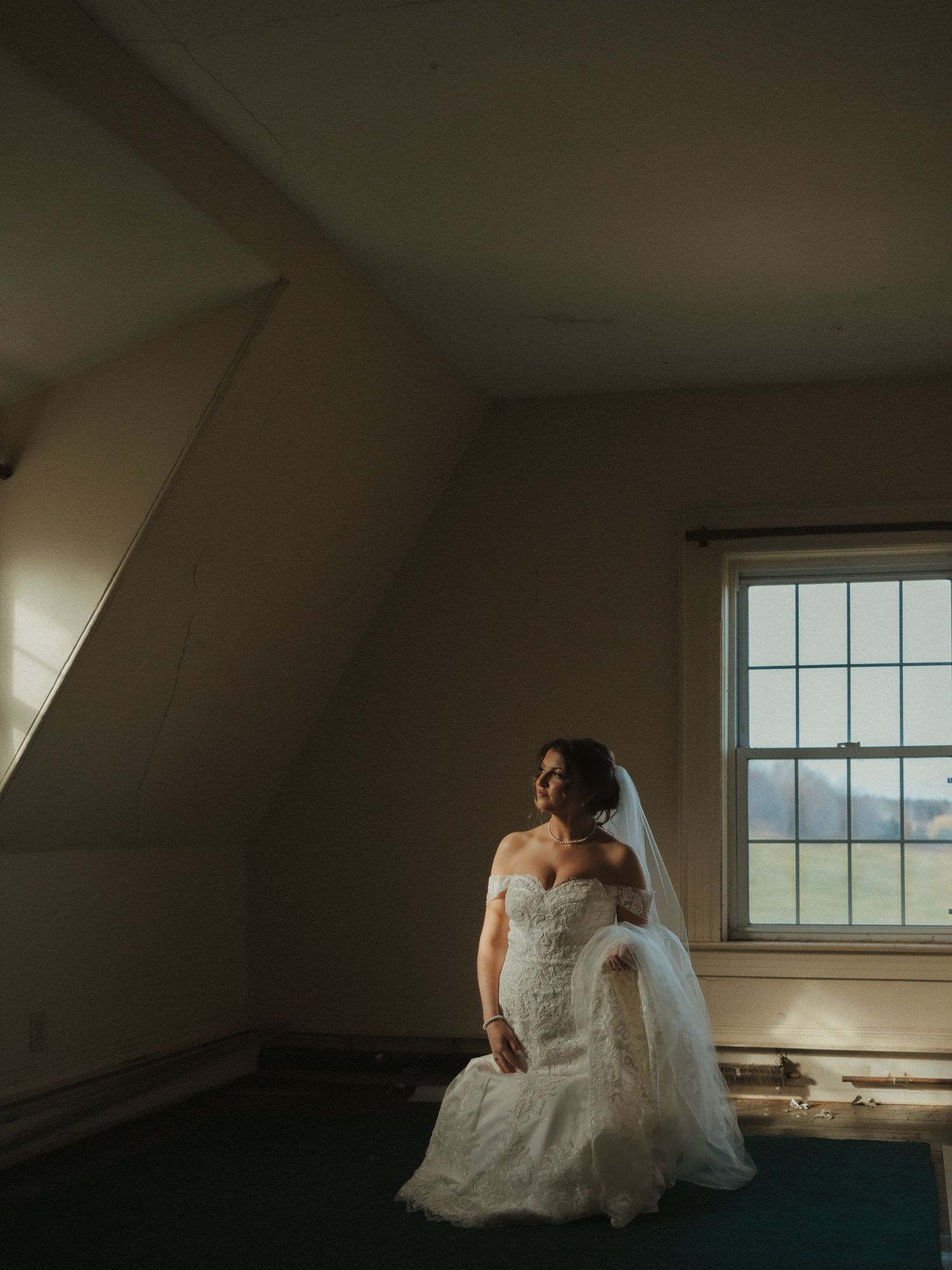 A bride in a white wedding dress sits contemplatively by a window in a sunlit attic room.