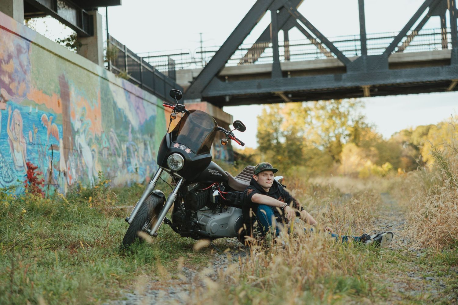 A motorcycle rests against graffiti-covered wall under a railway bridge surrounded by autumn foliage.