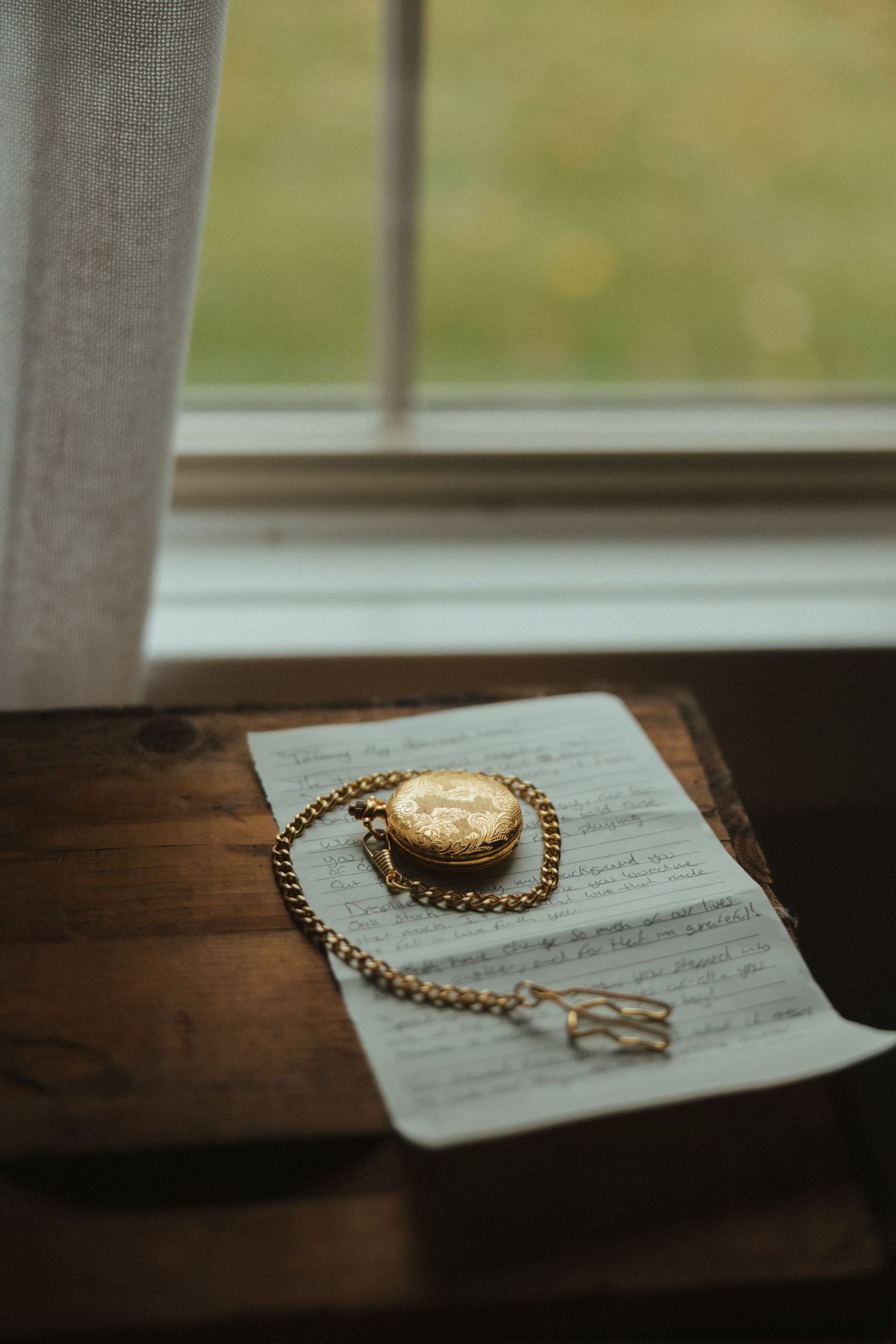 A gold necklace and ring rest on a handwritten paper note near a window ledge in soft natural light.