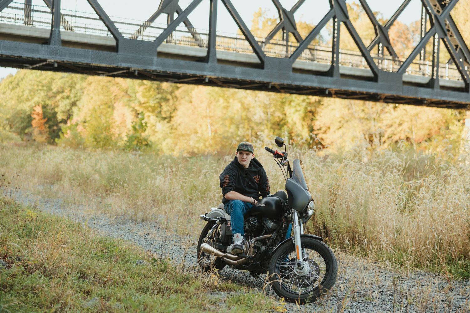 A motorcycle rider parked beneath a steel railway bridge on a sunny autumn afternoon.