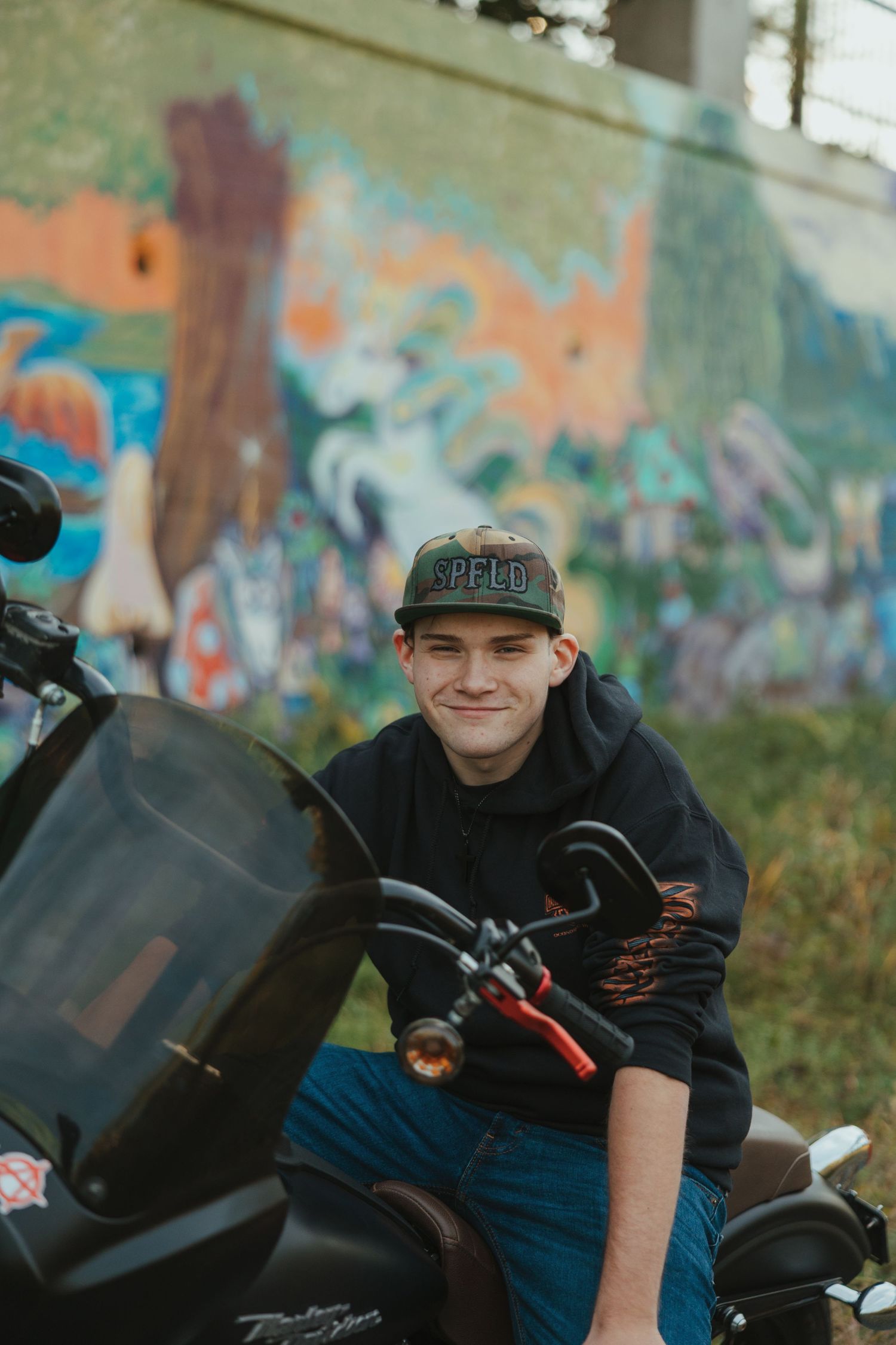 A motorcyclist in black clothing leans over a sport bike against a colorful graffiti wall background.