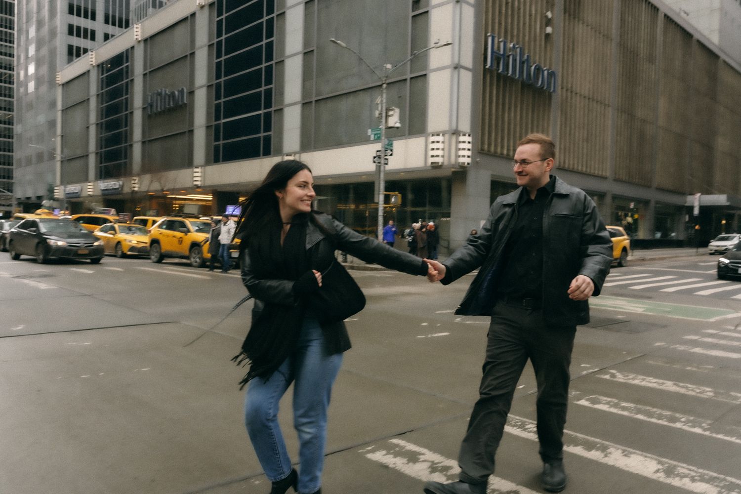 Couple holding hands and running across a busy New York City street near the Hilton hotel with yellow taxis.