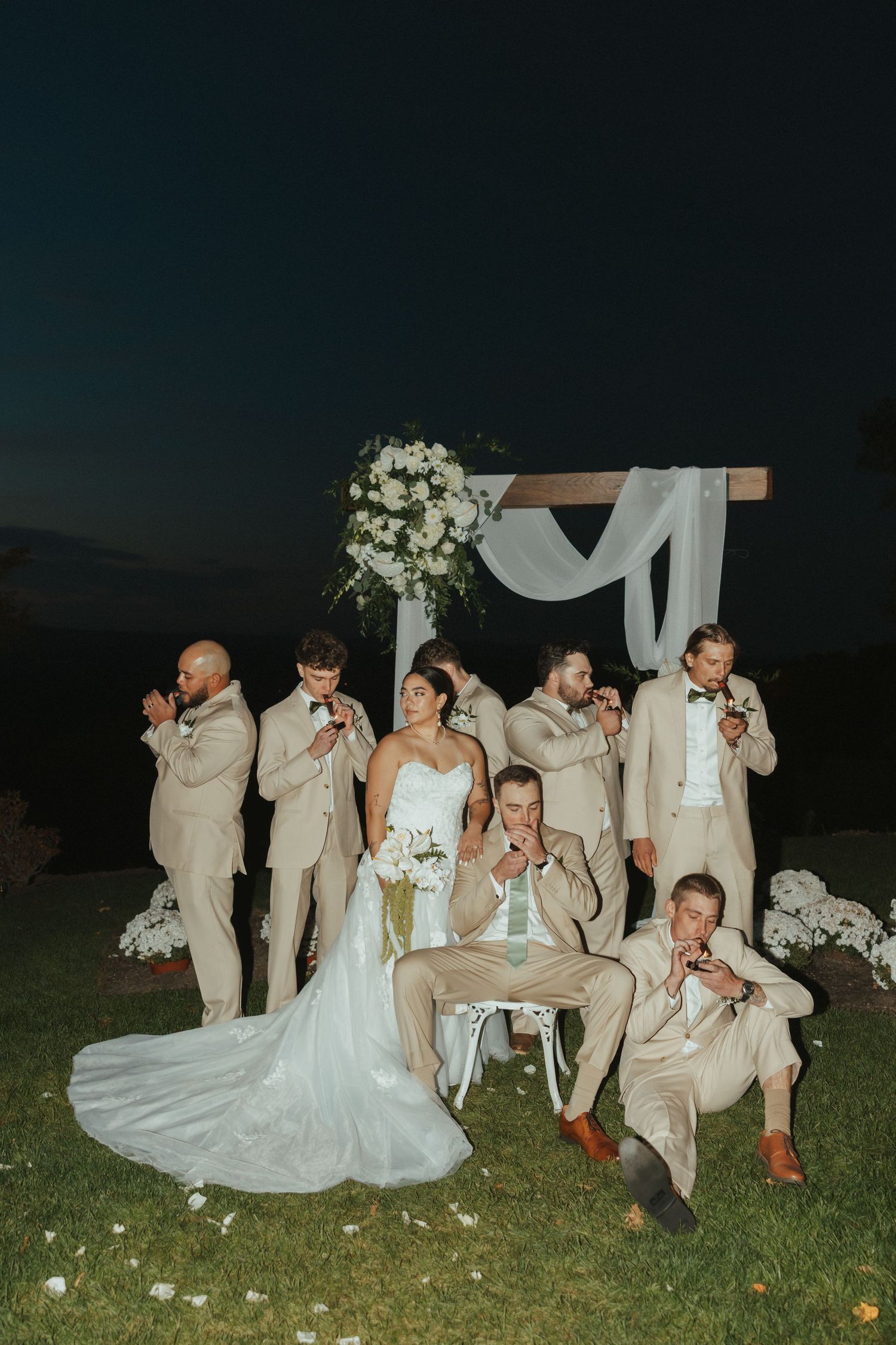 Wedding party in beige suits and white dress pose playfully on grass during outdoor evening celebration.
