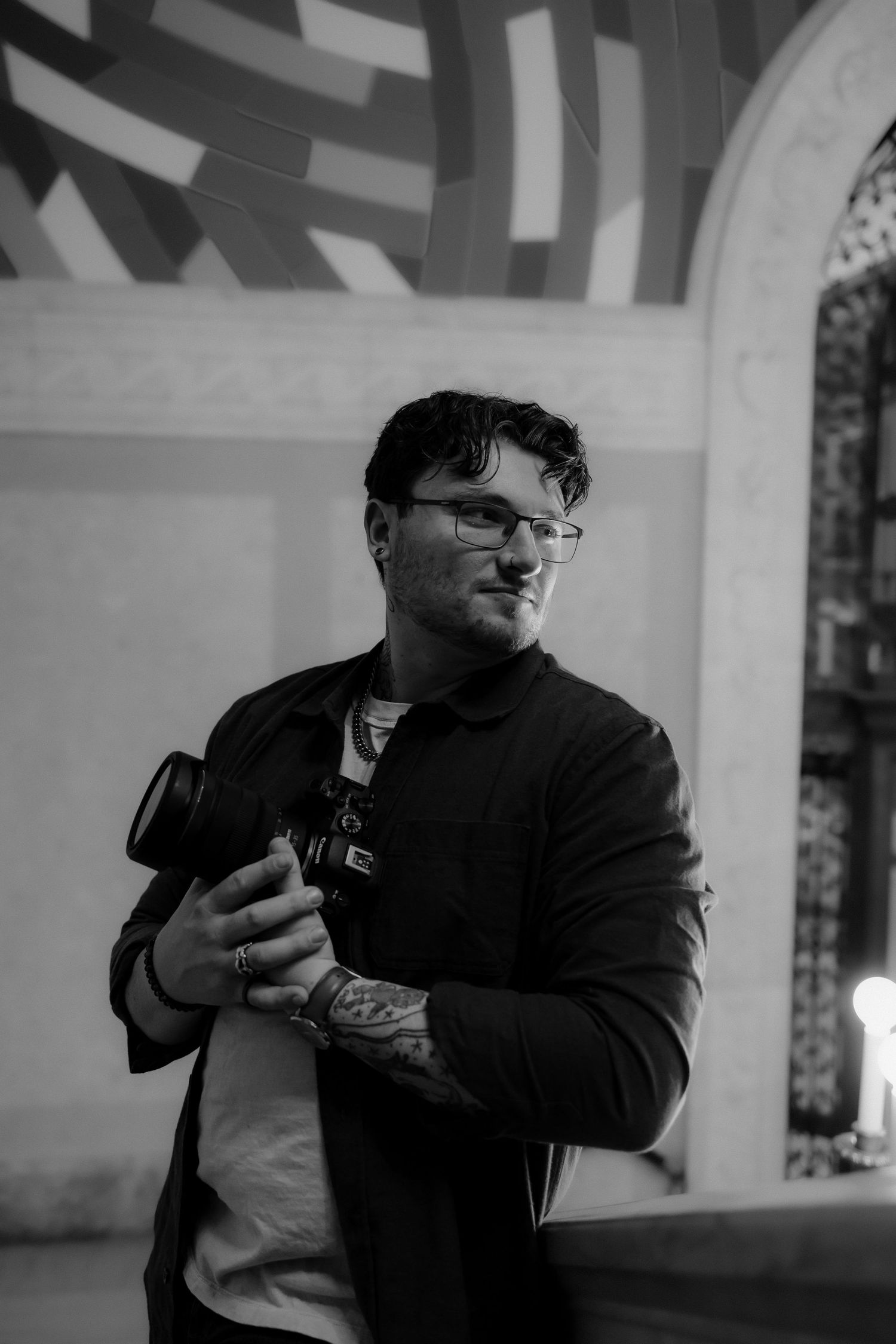 Black and white photo of a tattooed man with glasses holding a camera indoors.