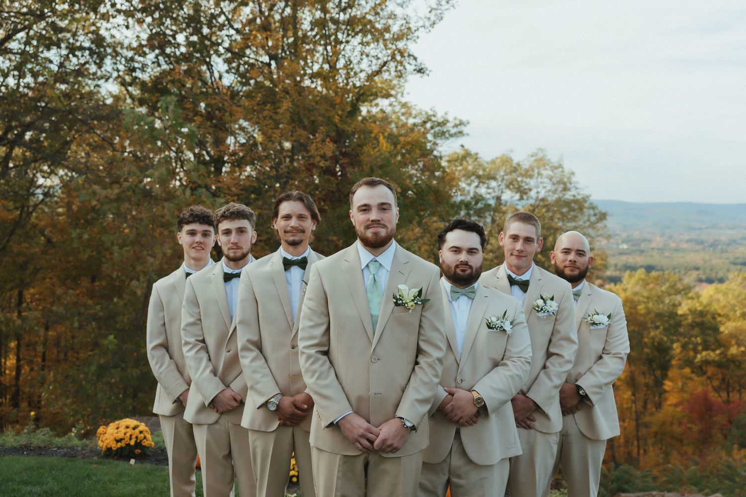 A wedding party in beige suits poses together on a hilltop during autumn with colorful fall foliage in the background.