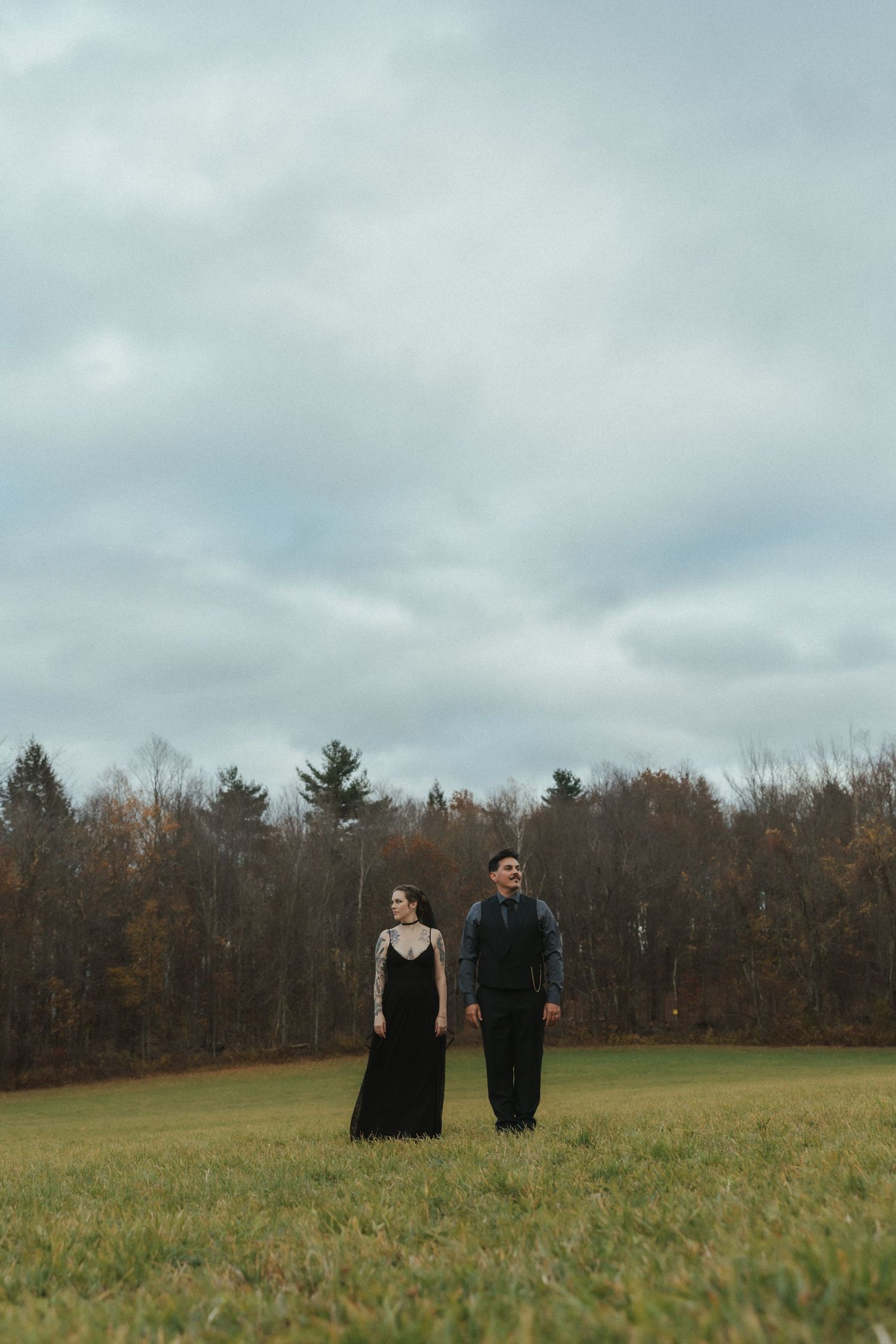 Couple standing in a field under cloudy skies wearing dark formal attire.