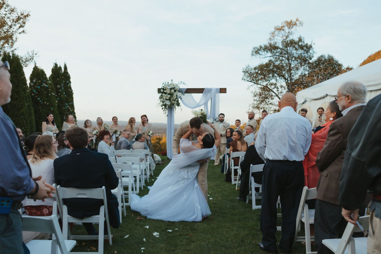 An outdoor wedding ceremony takes place on a grassy lawn with white chairs and guests gathered under blue skies.