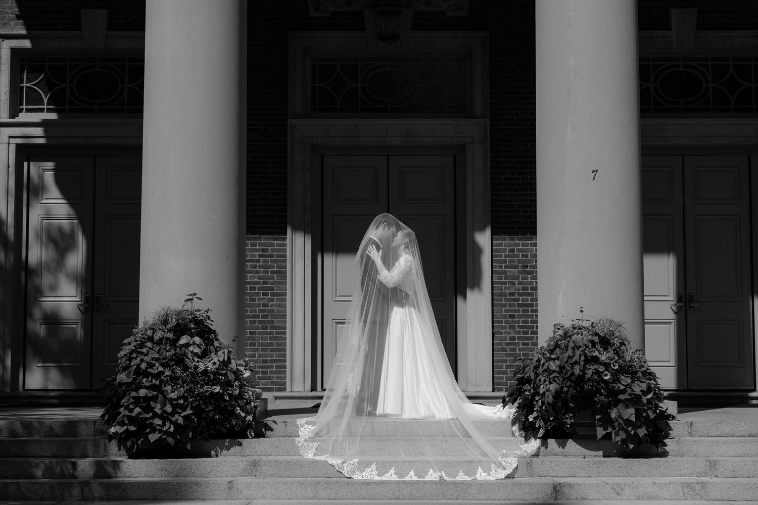 A wedding dress displayed in a window of a bridal shop with white columns and brick exterior.