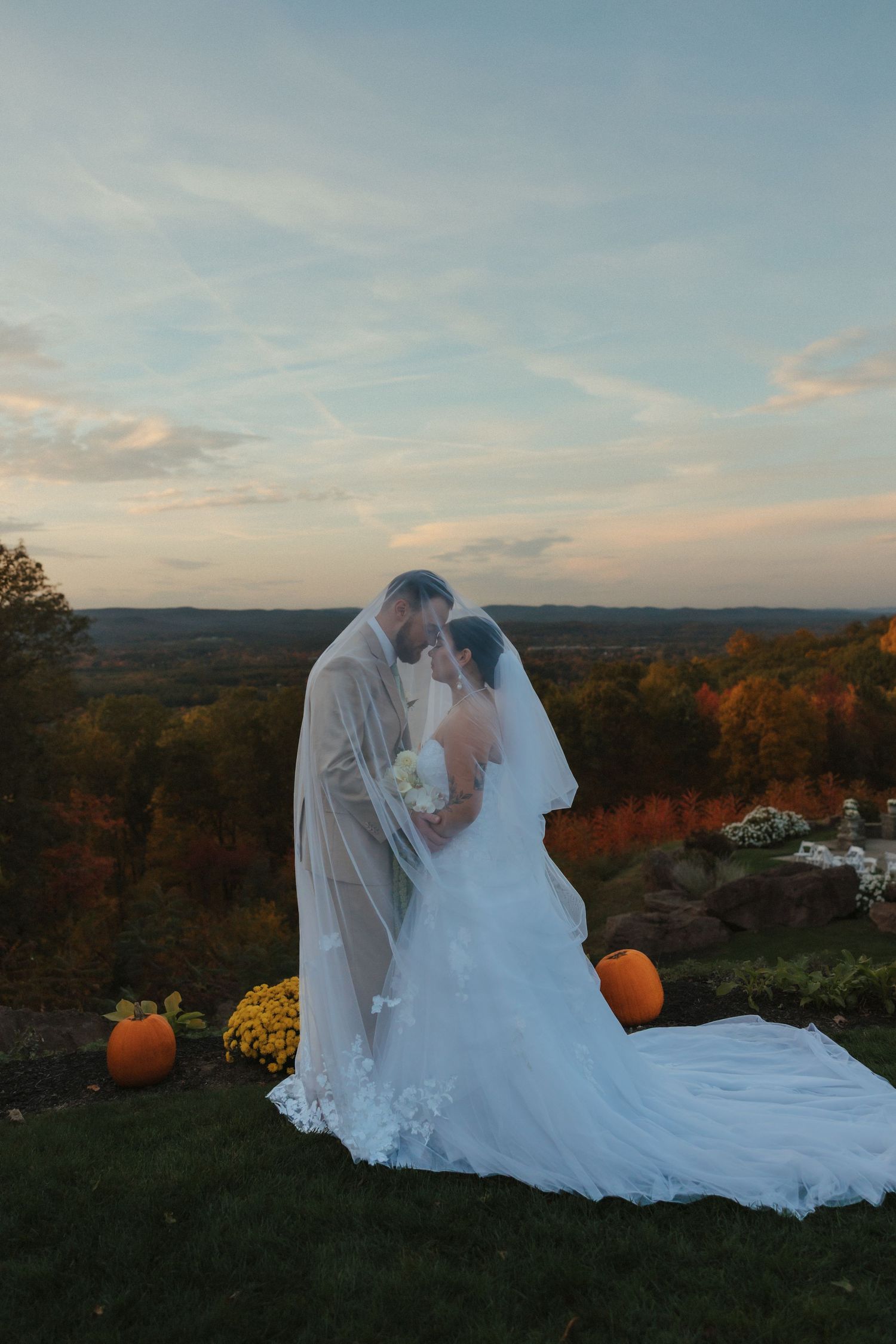 A romantic autumn wedding scene at sunset with a couple embracing on a hilltop decorated with pumpkins and fall flowers.