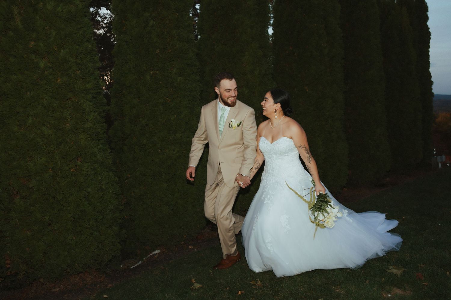 A bride and groom walk hand in hand along a hedge wall during their outdoor wedding.