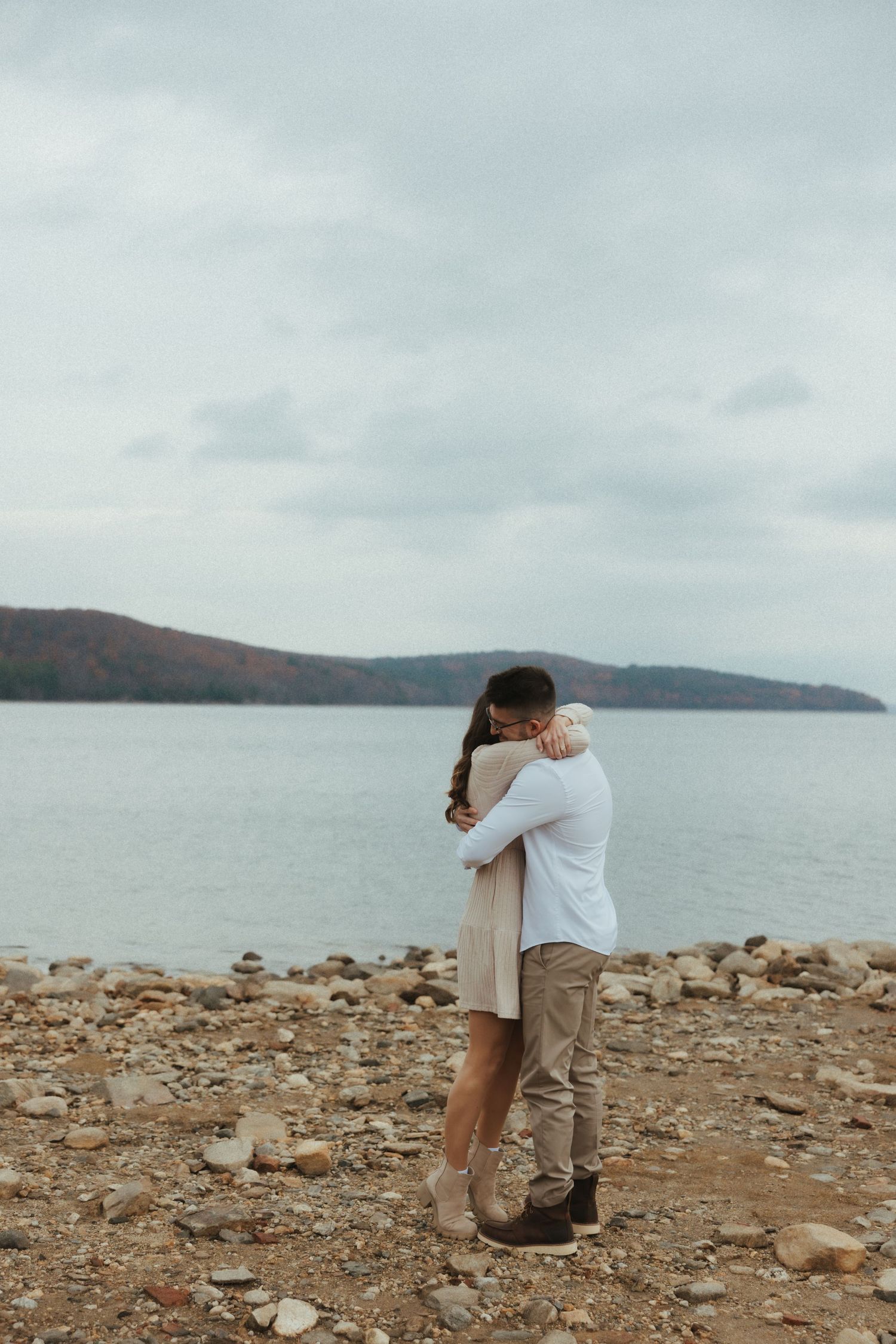A couple embraces on a rocky beach shoreline with mountains and cloudy skies in the background.