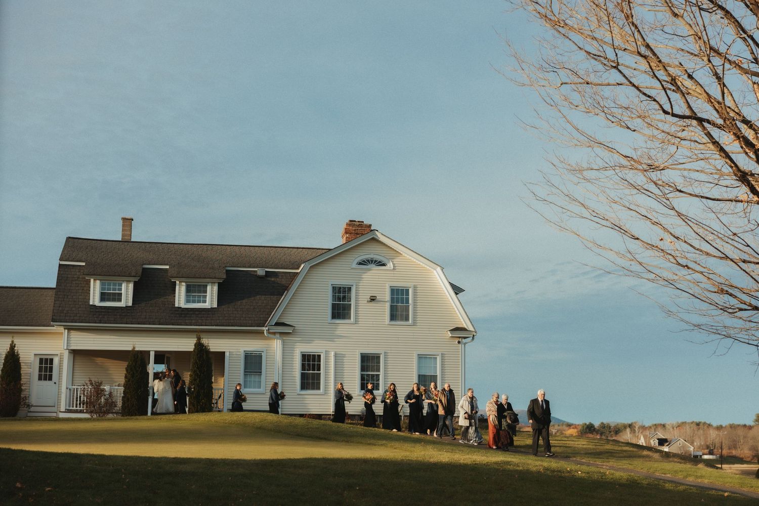 A panoramic view of a white farmhouse with dormers on a hilltop surrounded by people and bare winter trees.