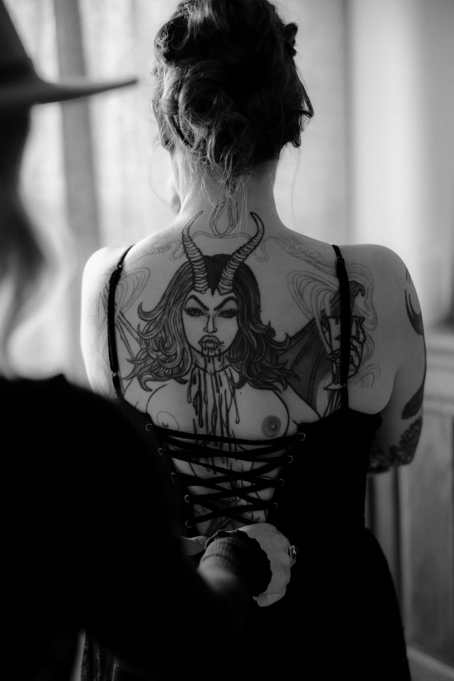 Four black and white photos show a woman's back with an intricate dark tattoo design spanning across her shoulders.