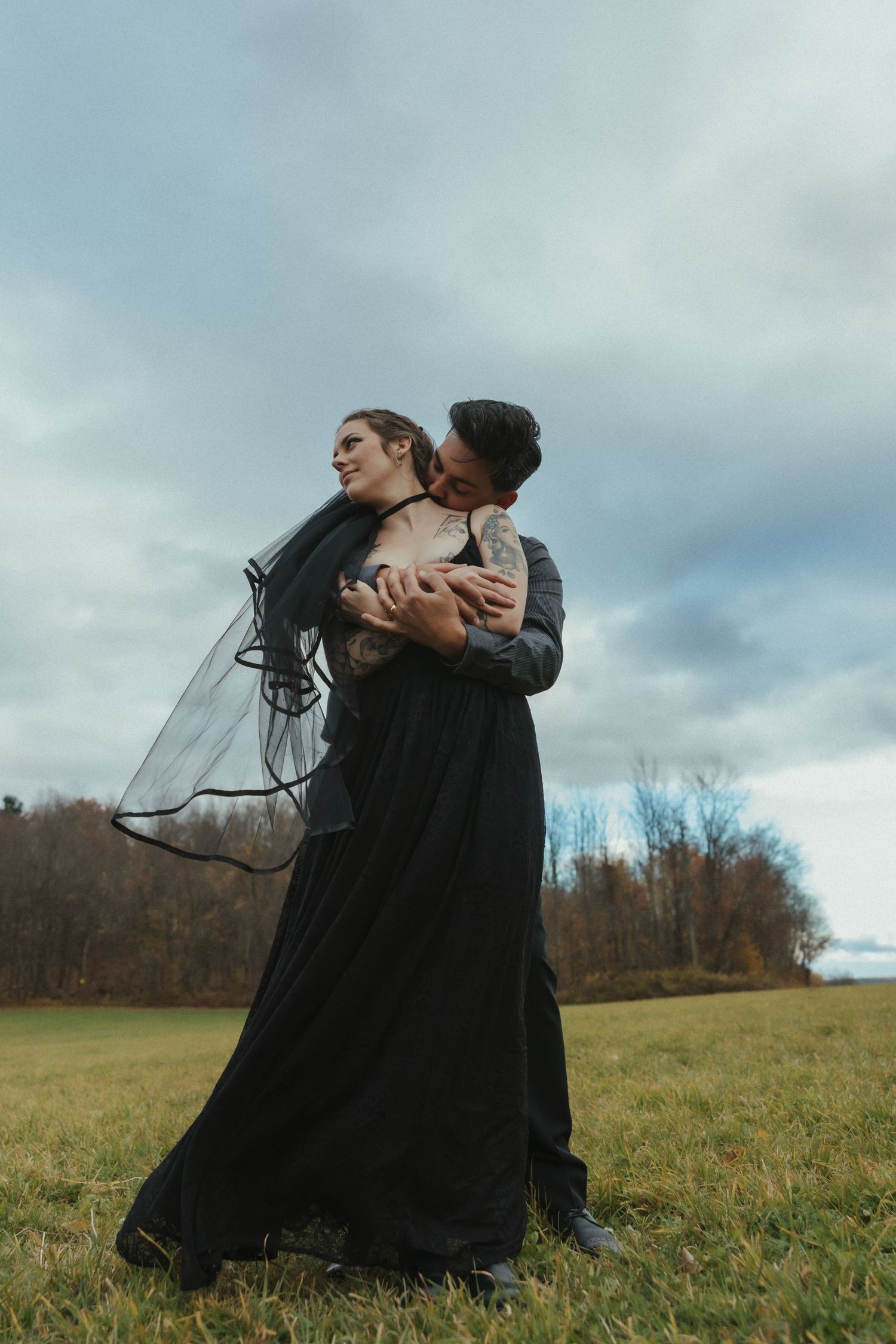 A couple in dark gothic attire embrace dramatically in a moody field under cloudy skies.