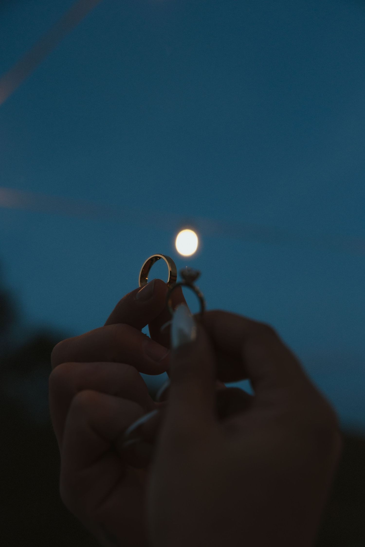 A series of hands holding up a ring against a dark blue twilight sky with the moon visible in the background.
