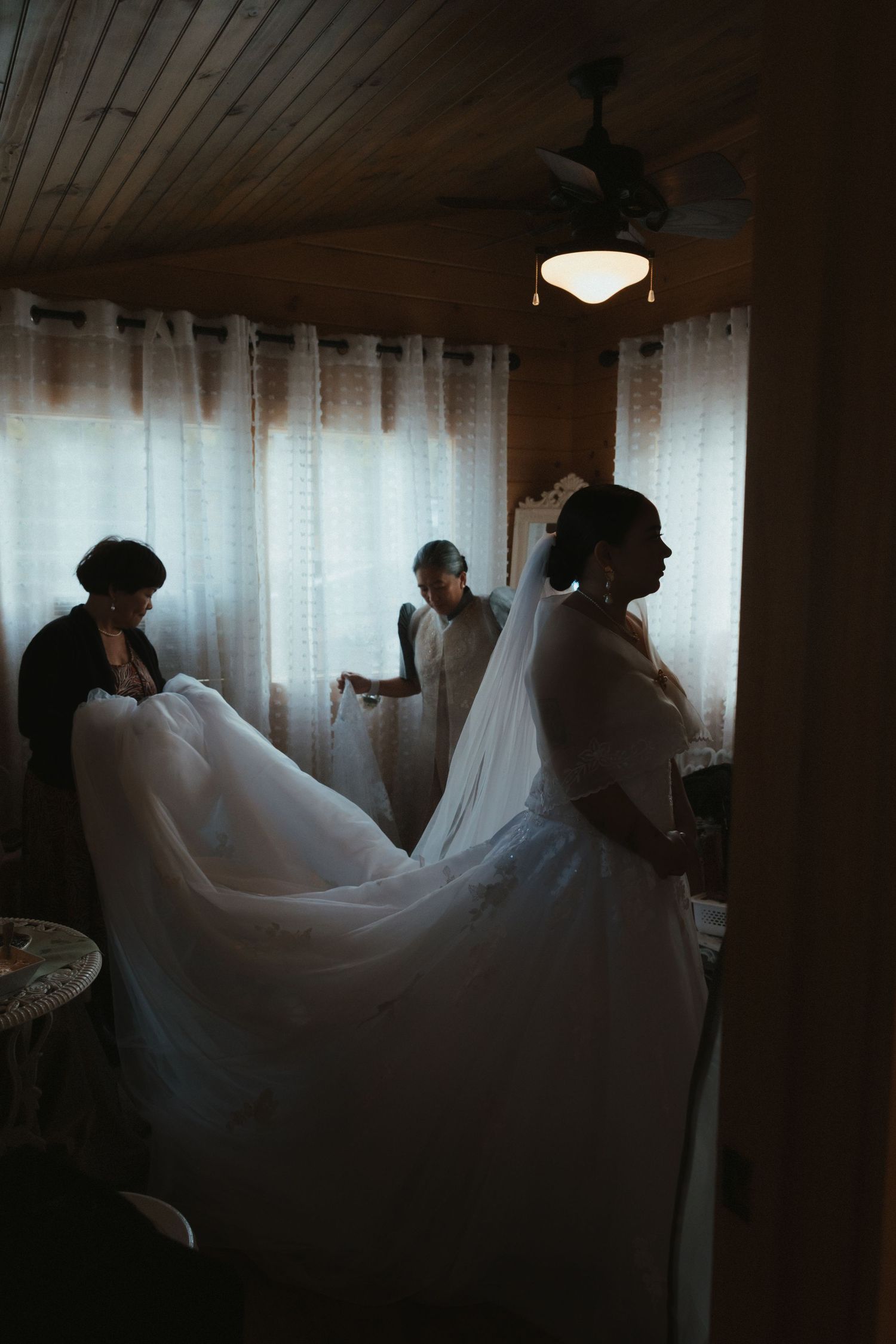 Flowing white wedding dresses create ethereal silhouettes against a backlit window in a dimly lit room.