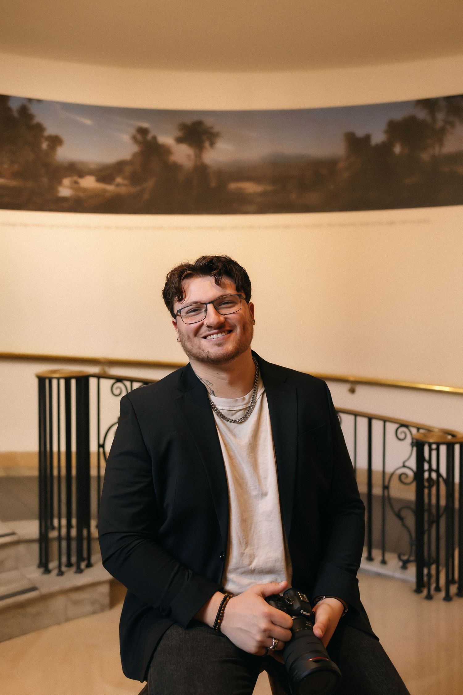 Smiling man in glasses and black blazer holding a camera, seated in an elegant hall with a landscape mural.