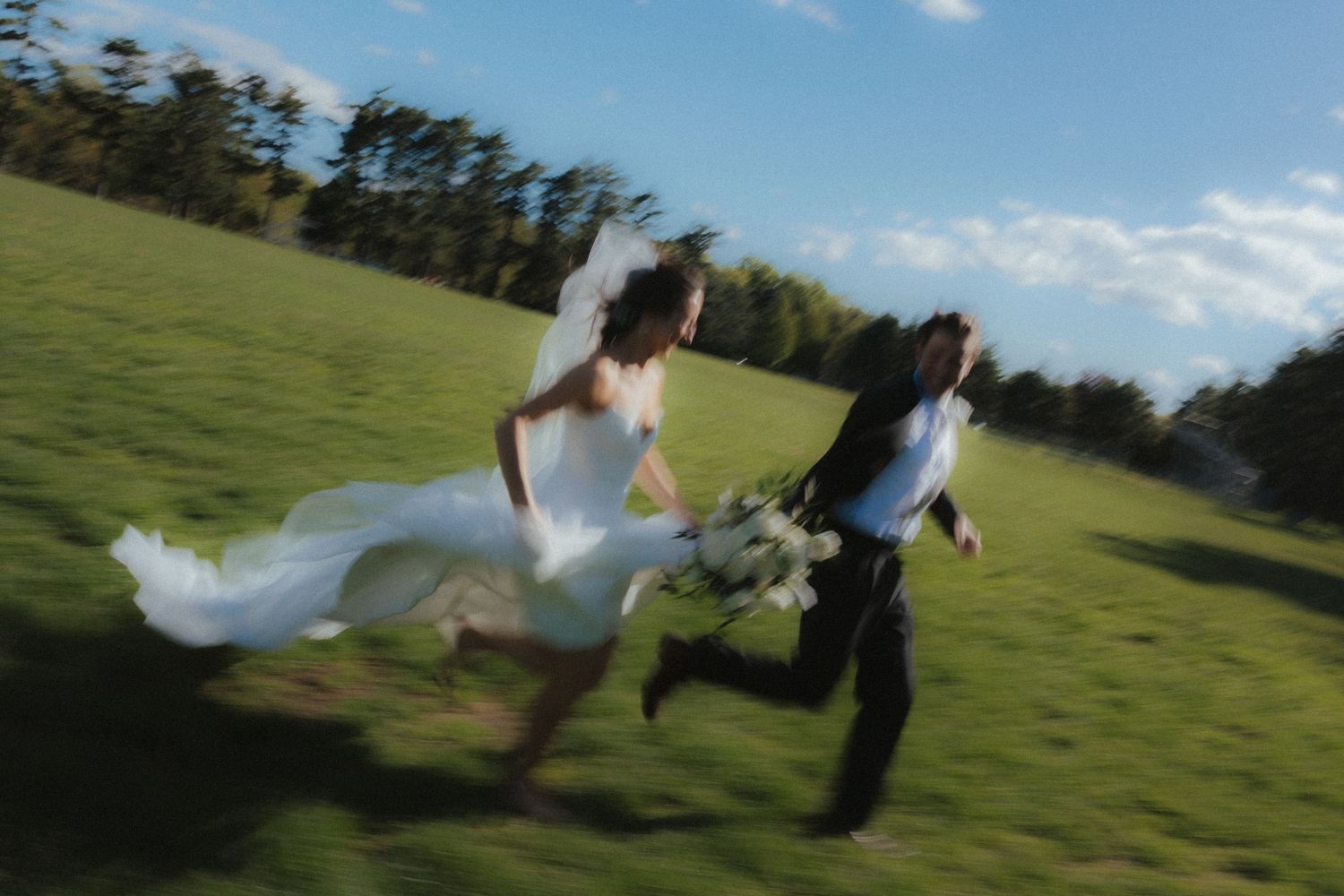Motion-blurred photo of a couple running together across a green field on their wedding day.